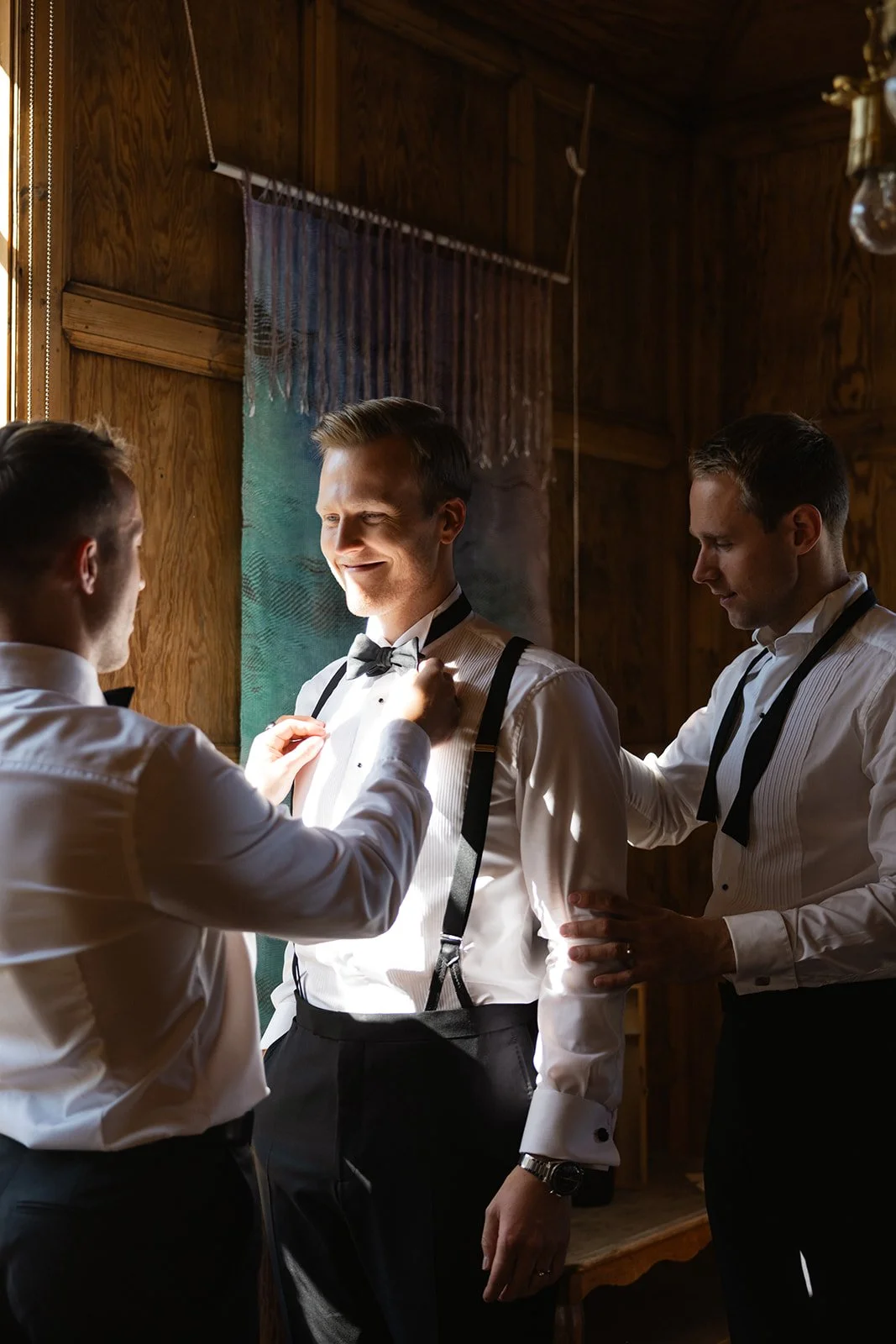 Tre menn i hvite skjorter og svarte bukser blir hjulpet med å ta på smoking og tilbehør av en kvinne.