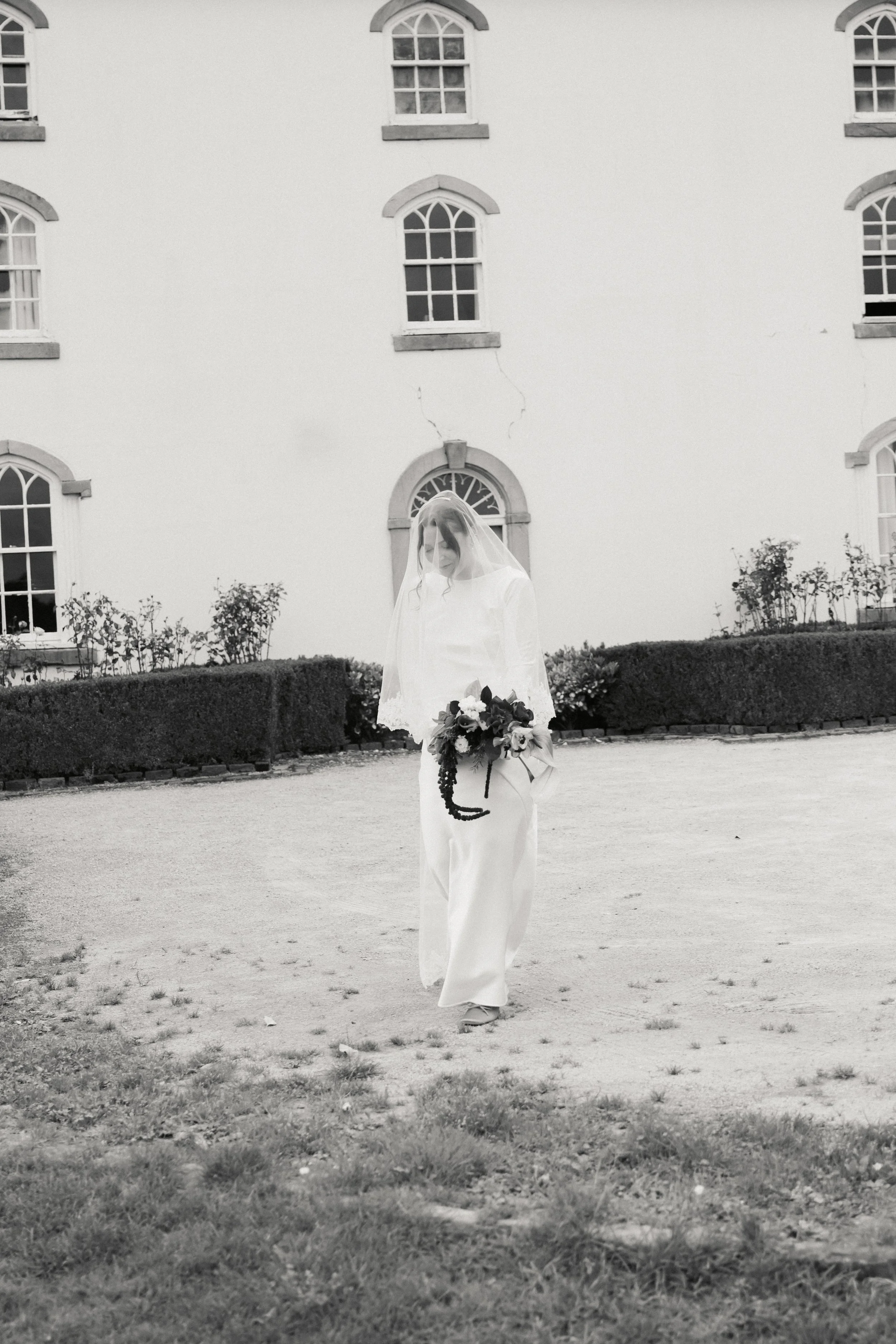 En kvinne i hvit brudekjole holder en blomsterbukett, står foran en hvit bygning med flere vinduer, omgitt av busker og en gresslette.