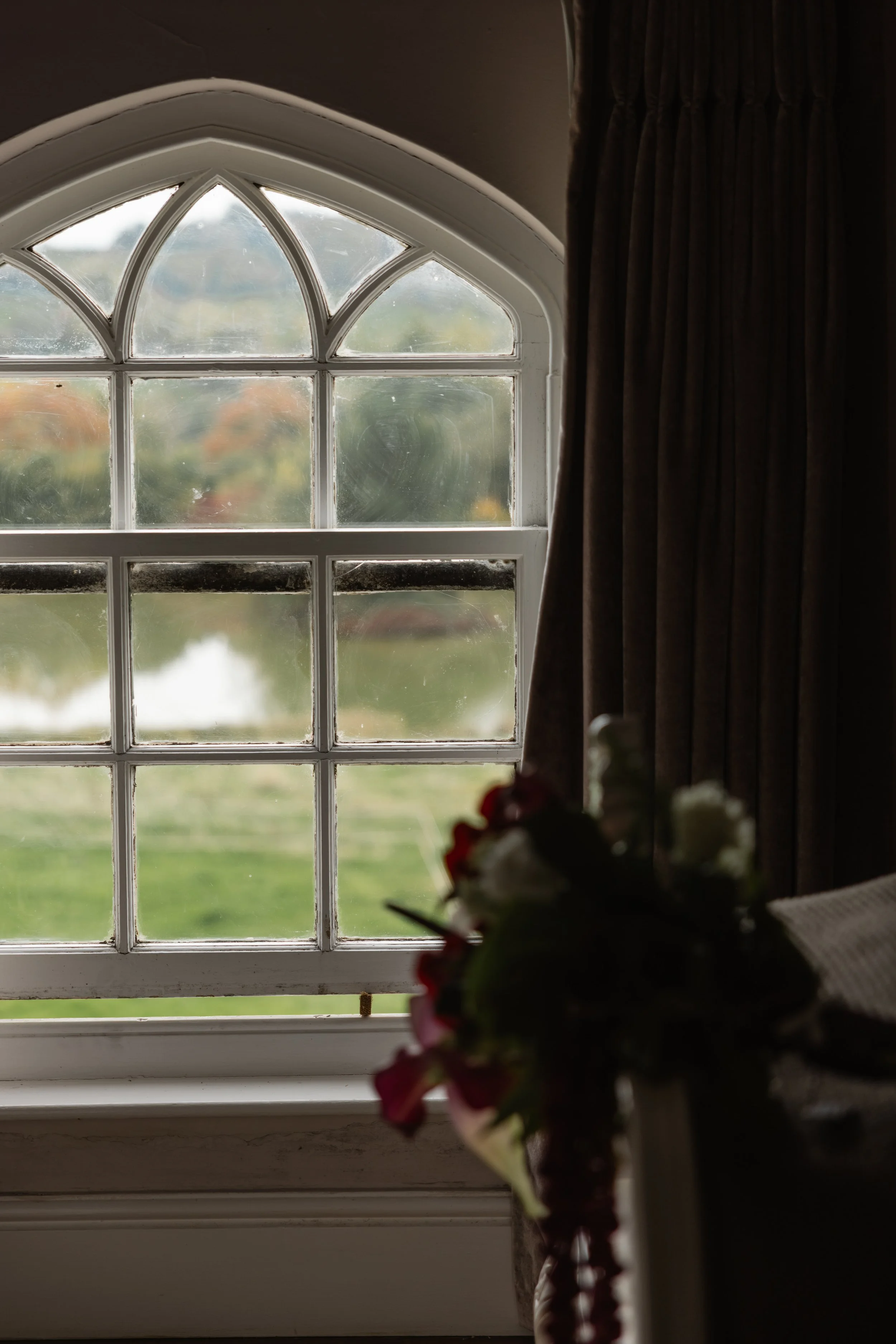 Et vindu med gammet hvit ramme og blyglass detaljer, sett fra innsiden av et rom, med mørke gardiner på siden. Utendørs er det en grønn landskap med vann og trær, litt ut av fokus.