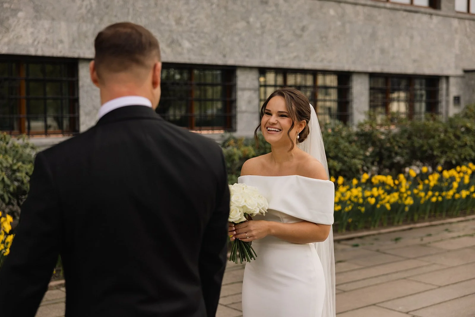 En brud med en hvit kjole og en brudebukett står foran en mann i en svart dress, smilende under et bryllup.