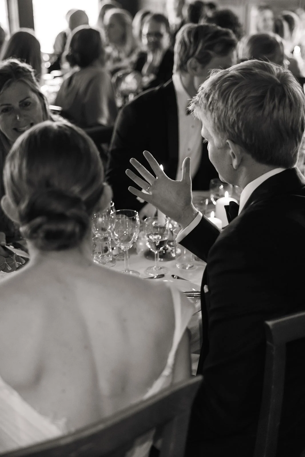 Svart-hvitt bilde av et middagsbord med mennesker i formelle klær, hvor en mann i dress snakker og gestikulerer mens en kvinne med oppsatt hår og kjole sitter med ryggen mot kamera.