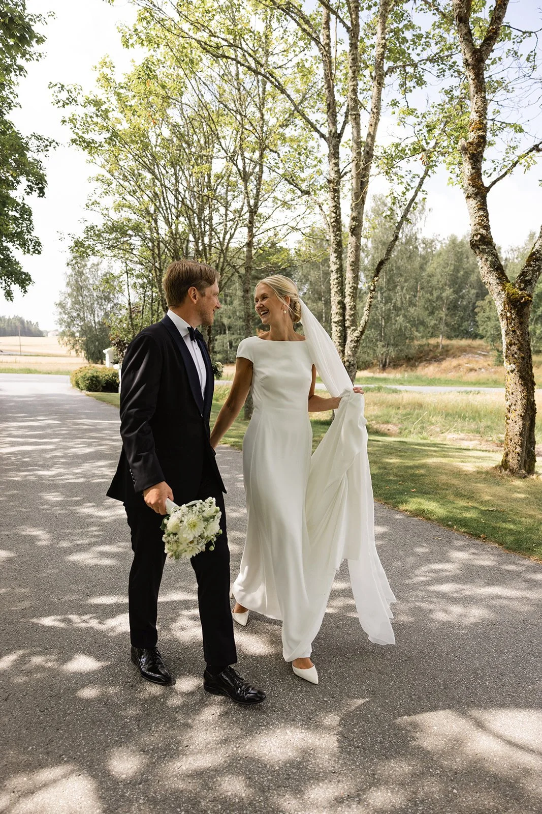 En lykkelig brud og brudgom går hånd i hånd på en landevei, omgitt av trær og grønt landskap, under en overskyet himmel, i en naturlig utendørs bryllupssetting.