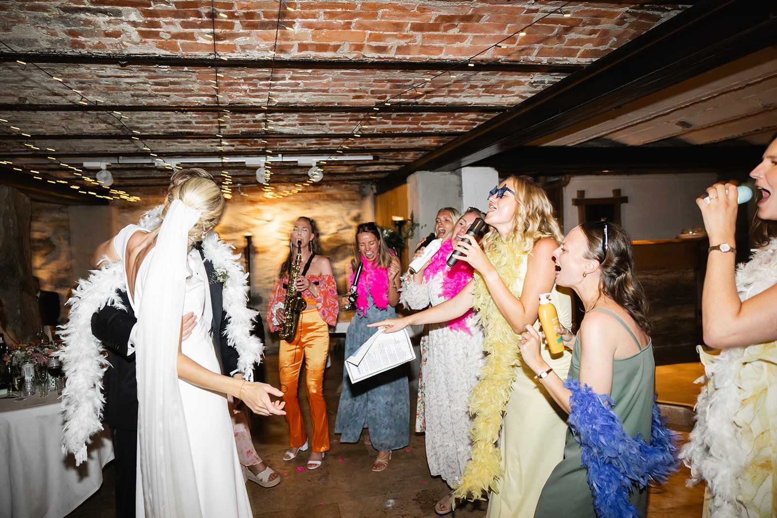 En gruppe kvinner og en mann feirer en bryllupsdans, kledd i fargerike klær og boas, i et treverk- og mursteinslokale med dekorasjoner og blomster.