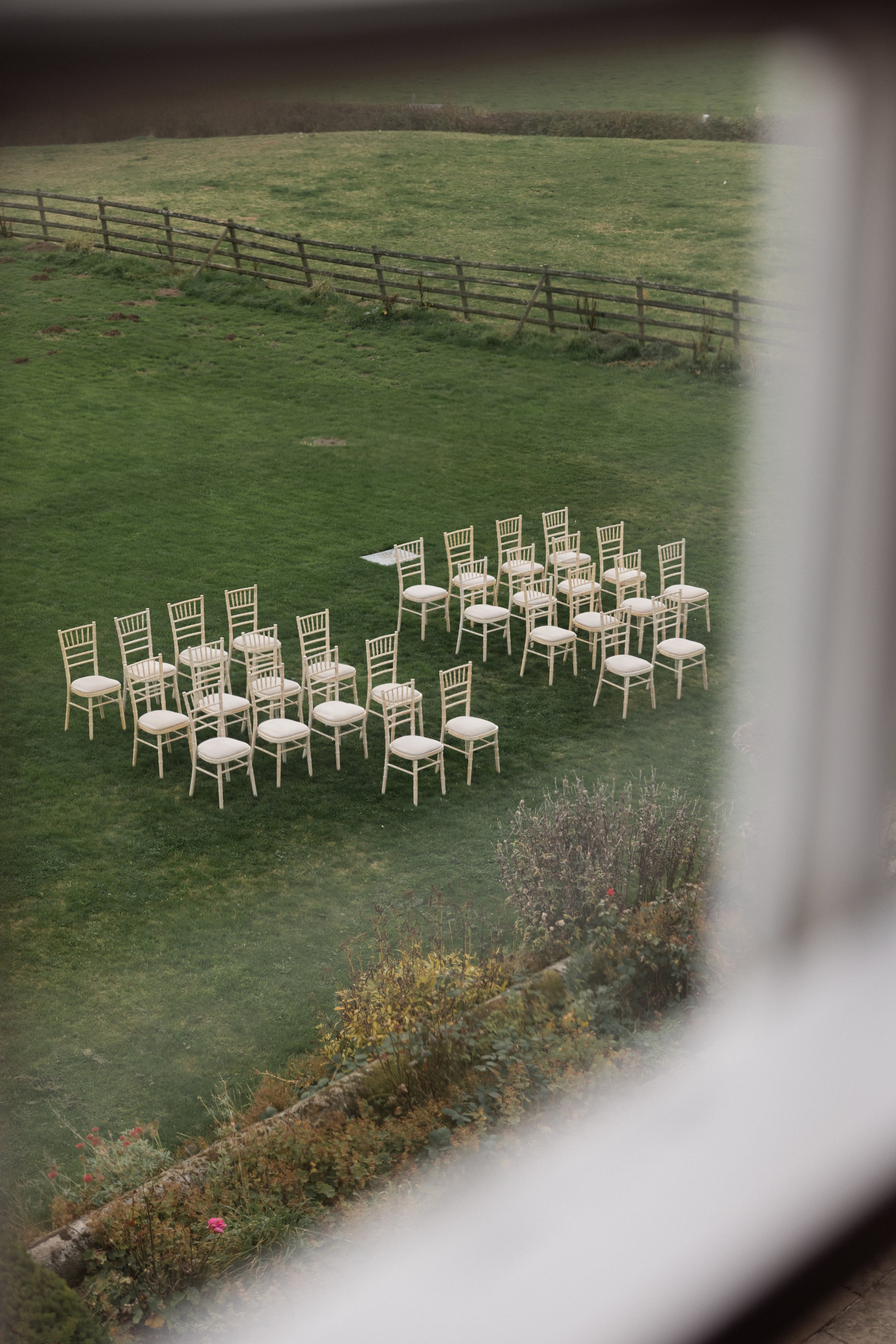 En gruppe hvite stoler er plassert på gresset i en hage, sett fra et vindu med gjennomsiktig glass.