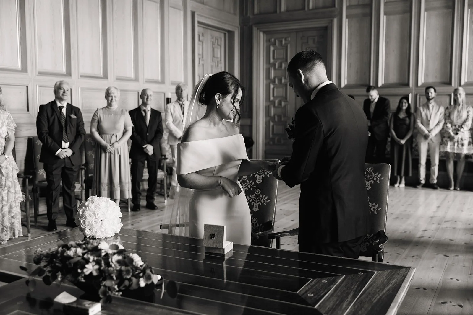 Et brudepar som gifter seg i et tradisjonelt rom med trevegger, hvor bruden og brudgommen står foran hverandre og holder hendene, omgitt av familie og venner.