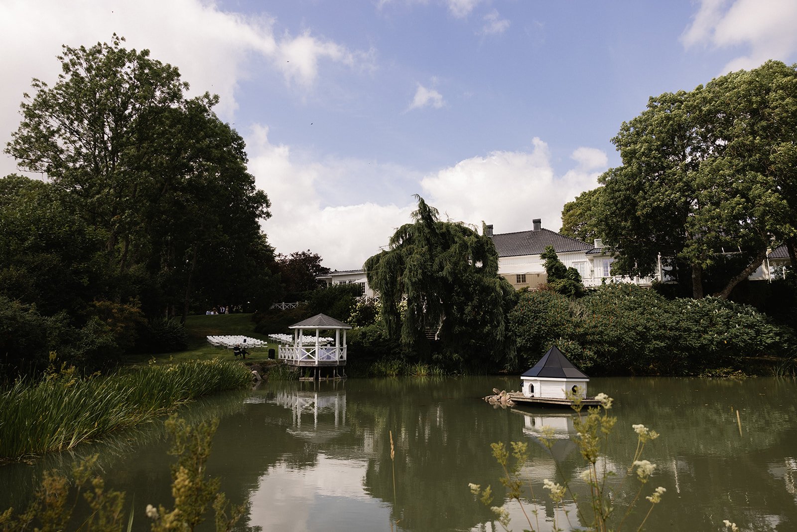 Bilde av en dam omgitt av trær og busker med en liten lystbåthytte og en terrasse i bakgrunnen.