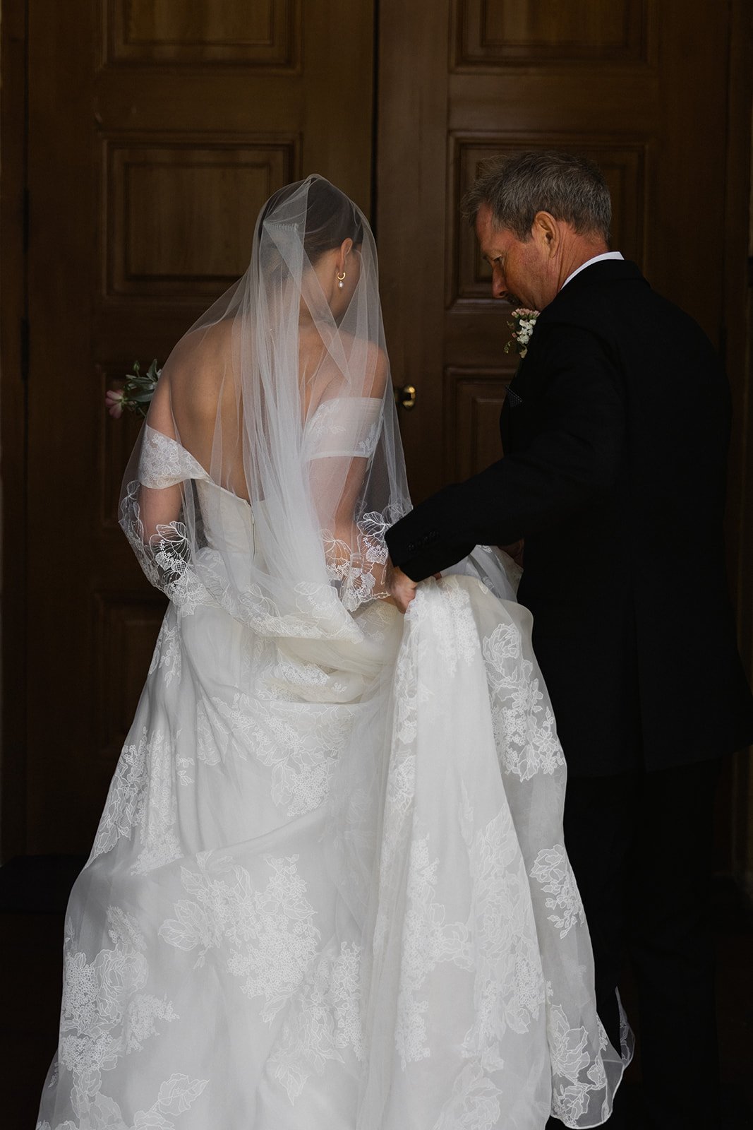 En brud i hvit brudekjole og en mann i svart dress som hjelper henne med å bære brudekjolen, foran en trepanelvegg.