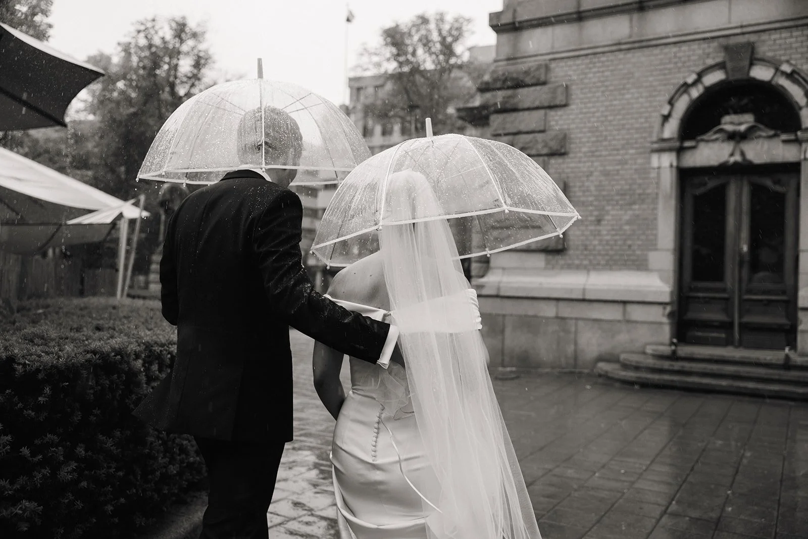 Bryllupsparet går i regnet med transparent paraplyer, kvinnen har brudekjole og brudeblonder, mannen har dress, foran et gammelt mursteinsbyggverk.