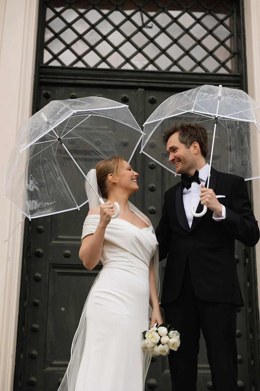 To bryllupskledd personer, en kvinne med hvit kjole og en mann i tuxedo, står under klare transparent paraplyer foran en stor, mørk port.