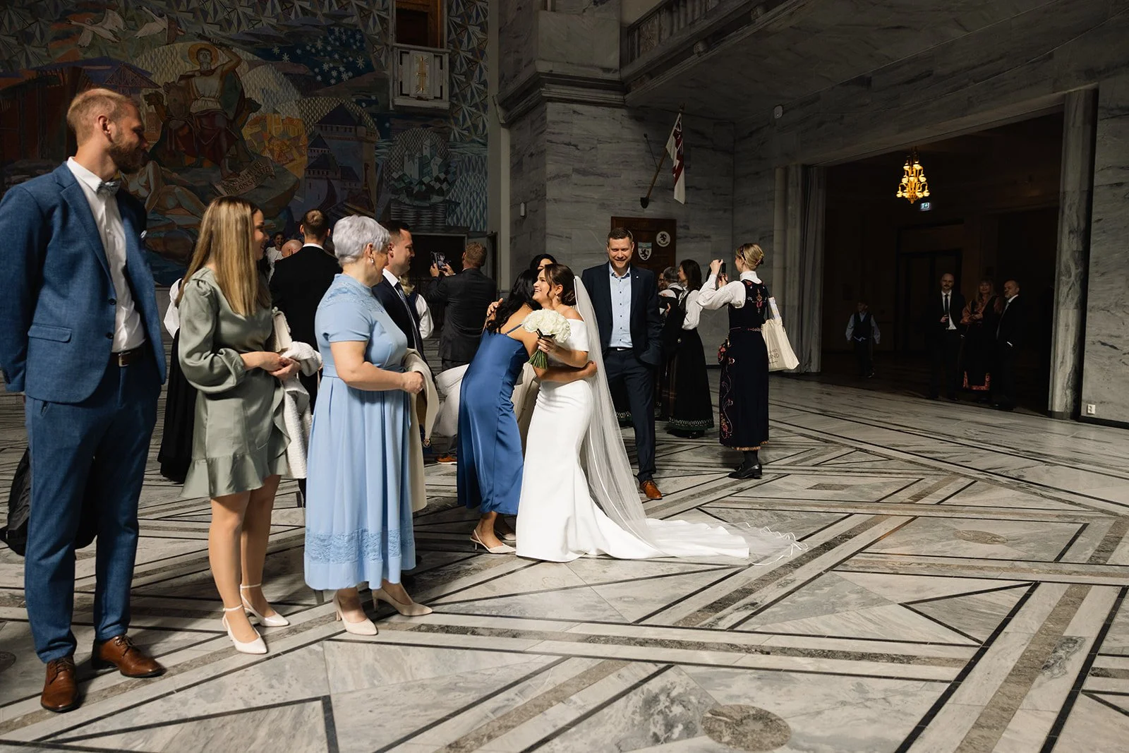 En gruppe mennesker på en vielsesceremoni med en brud i hvitt og en gjest i en blå kjole som klemmer. Andre gjester står i rekker og filmer eller observerer. Innendørs med marmorgulv, kunst på veggen og en gammel sjekk-lysekrone.