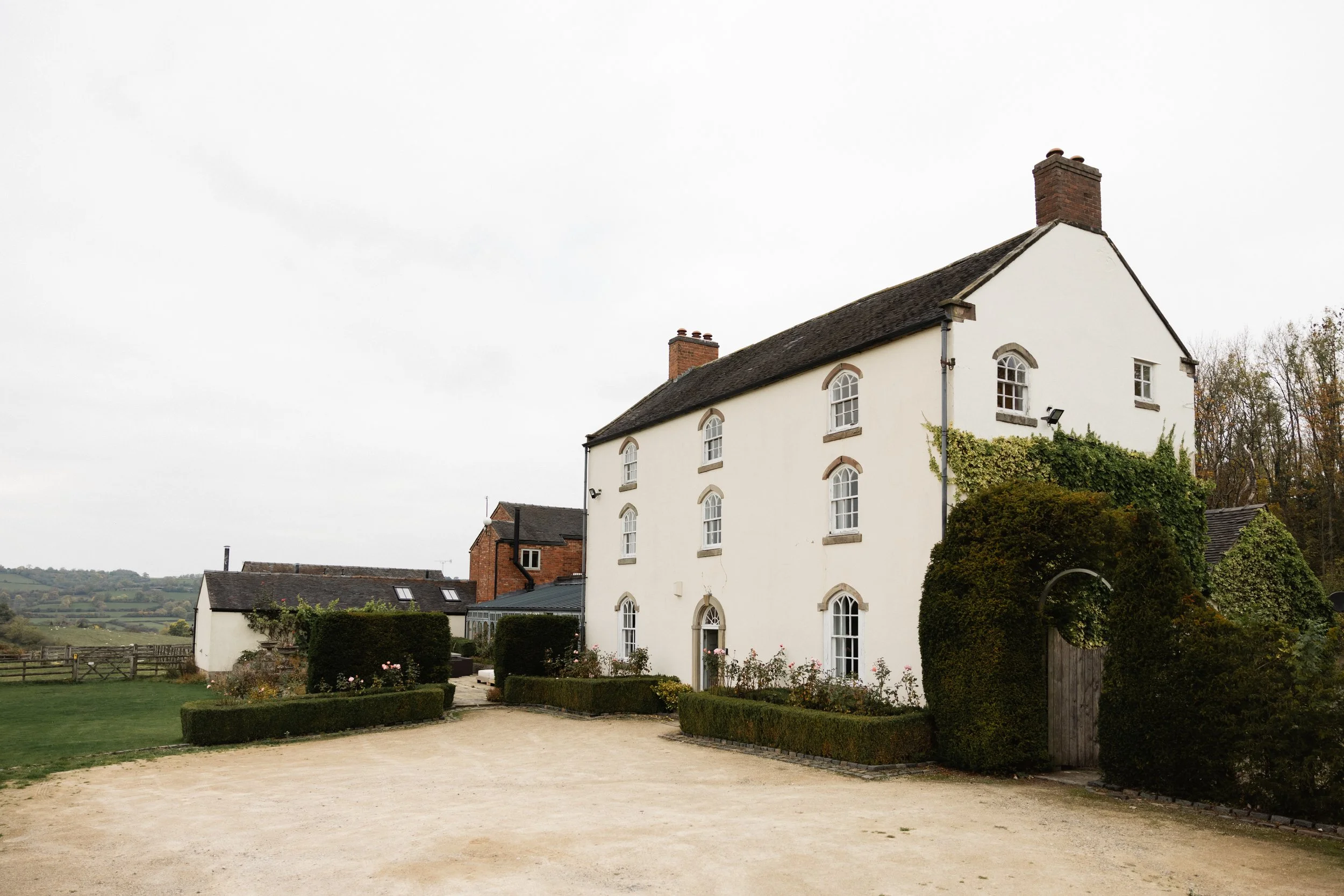 Hvitt hus med flere etasjer, omgitt av hagedense busker og blomster, med en gruslagt vei foran og en åpen landskap i bakgrunnen