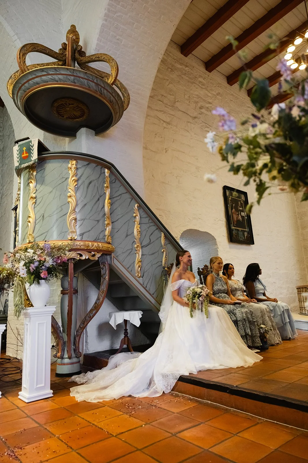 En bryllupshvit kjole med brudepike eller brud som sitter ved siden av fire kvinner i ulike kjoler i en kirke. Det er blomsterdekorasjoner og en figur av en kongekrone på veggen.