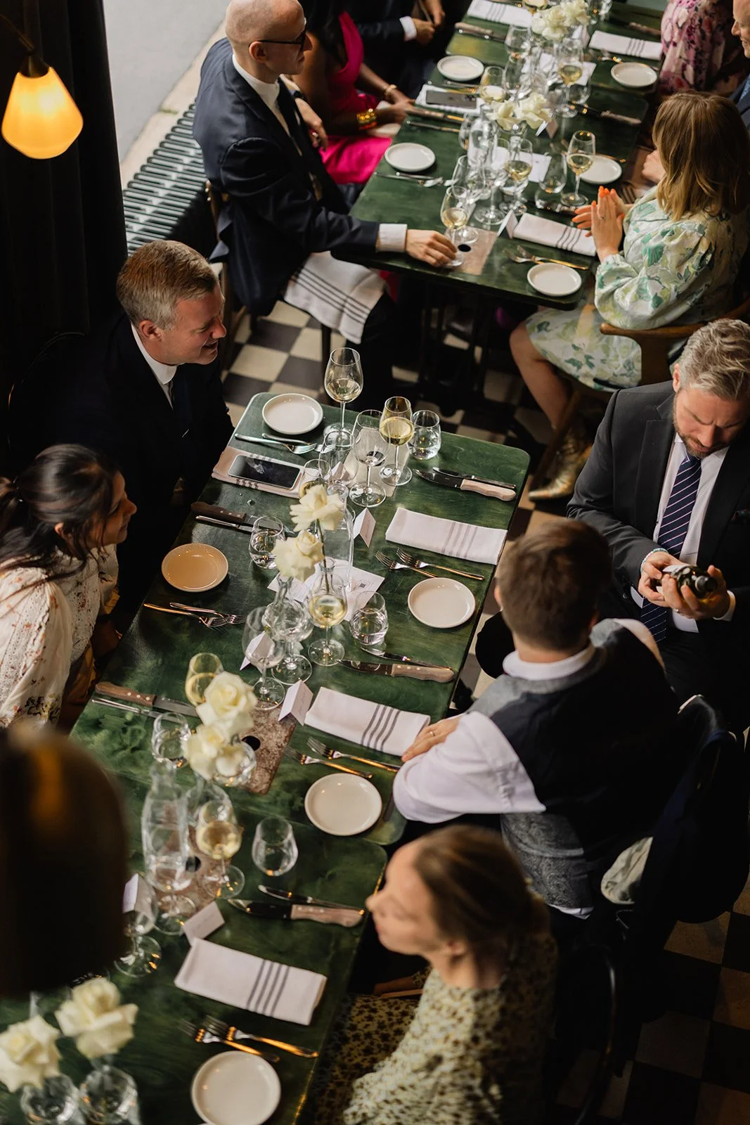 Middagsselskap rundt lang bord med hvite blomster, hvite tallerkener, sverd, vinglass og servietter, i et elegant innendørs miljø.