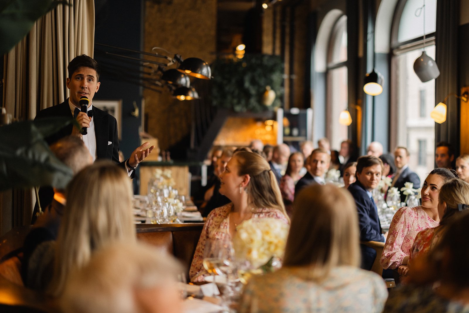 En mann i tuxedo holder en mikrofon og snakker foran en gruppe mennesker som sitter ved et langbord i et vakkert innendørs sted, sannsynligvis under en begivenhet eller seremoni.