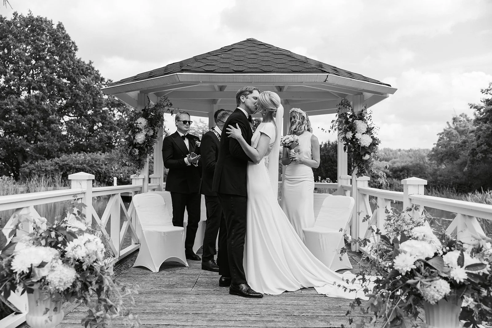 Et bryllupsceremoni i utendørs lysthus med brudepar som kysser, omgitt av brudepike og forlover, dekorert med blomster.