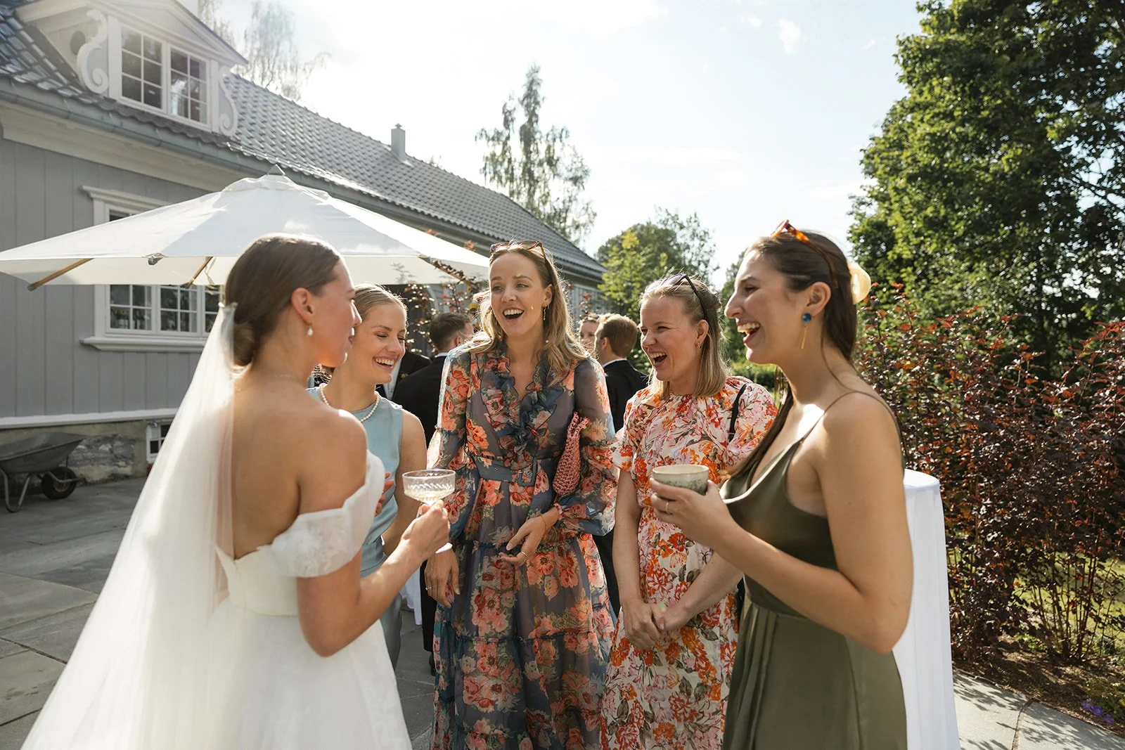 En gruppe mennesker som smiler og ler, deltar ved et utendørs bryllup eller fest, med en hage og en bygning i bakgrunnen.