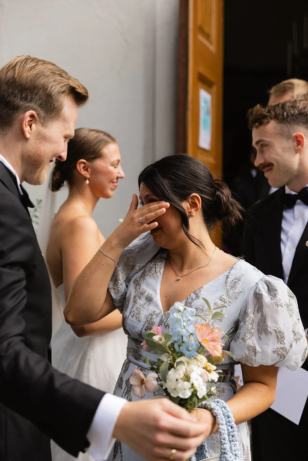 Emosjonell scene på en bryllupsfest med personer i formelle klær, der en kvinne i midten er rørt og gråter, mens andre rundt henne smiler og har det bra.
