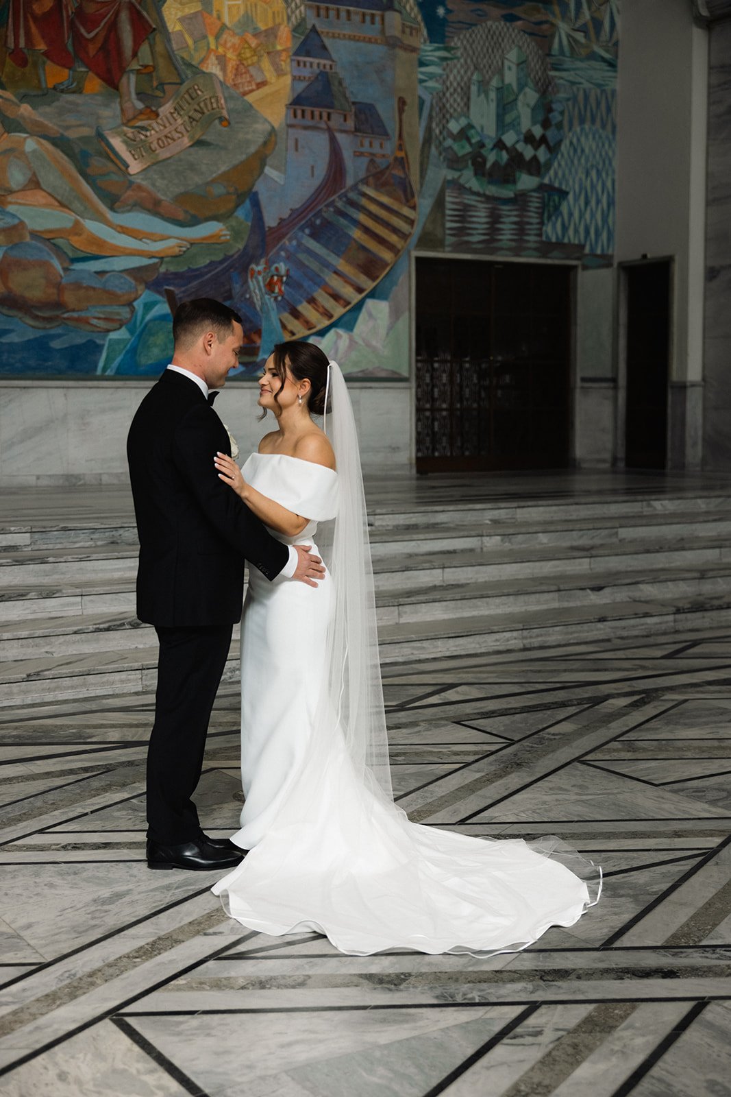 Et brudepar står og ser på hverandre i en kirke, med et fargerikt veggmaleri i bakgrunnen.