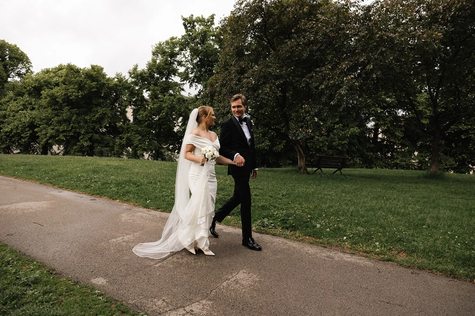 Et brudepar går hånd i hånd på en parkvei, omgitt av grønne trær, hvor kvinnen har på seg en hvit brudekjole med slør og holder en blomsterbukett, og mannen har på seg en svart Dress og slips.