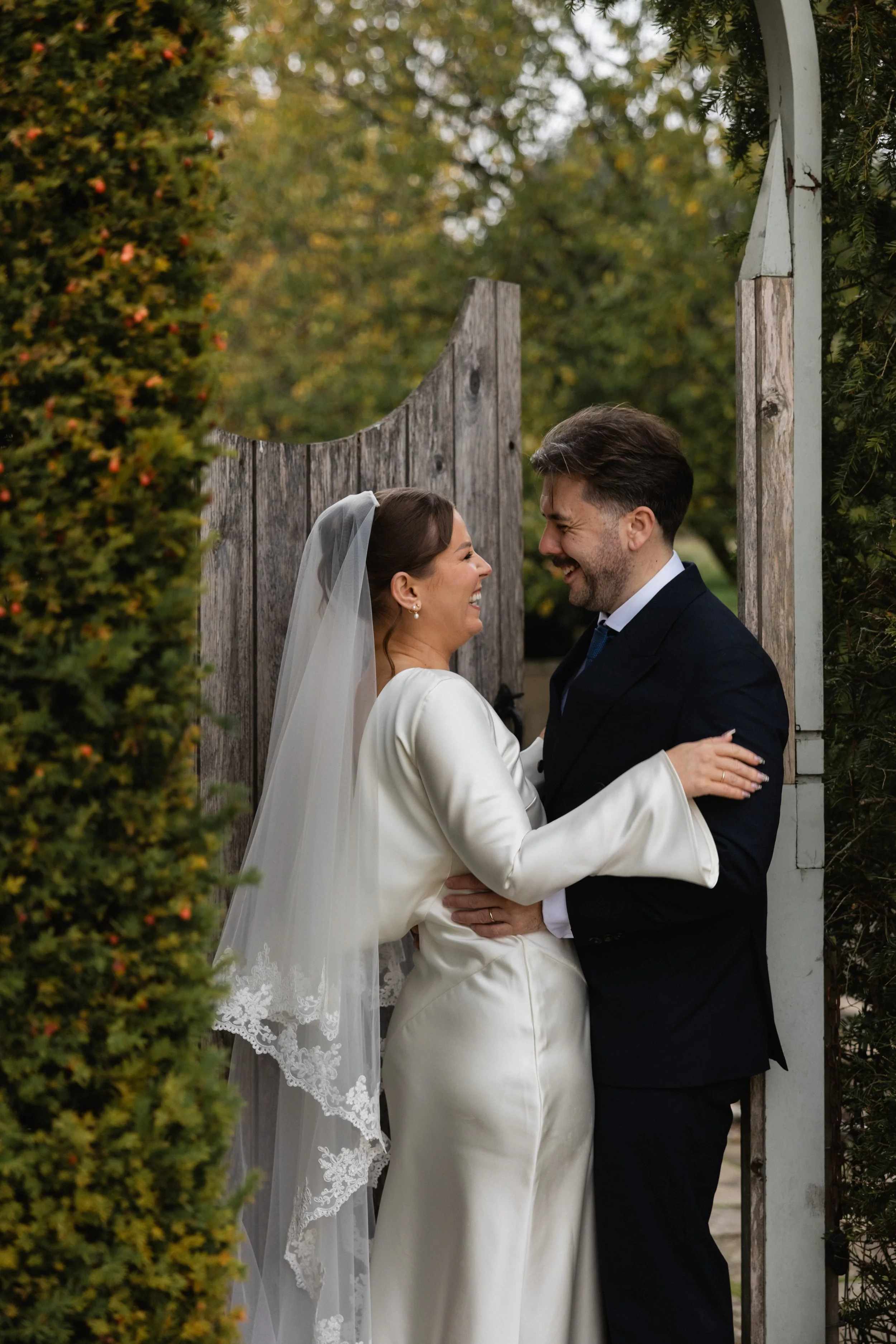 Et lykkelig bryllupspar som ler sammen, stående ved en treport omgitt av grønne busker og trær, i høstmiljø med varmt lys.