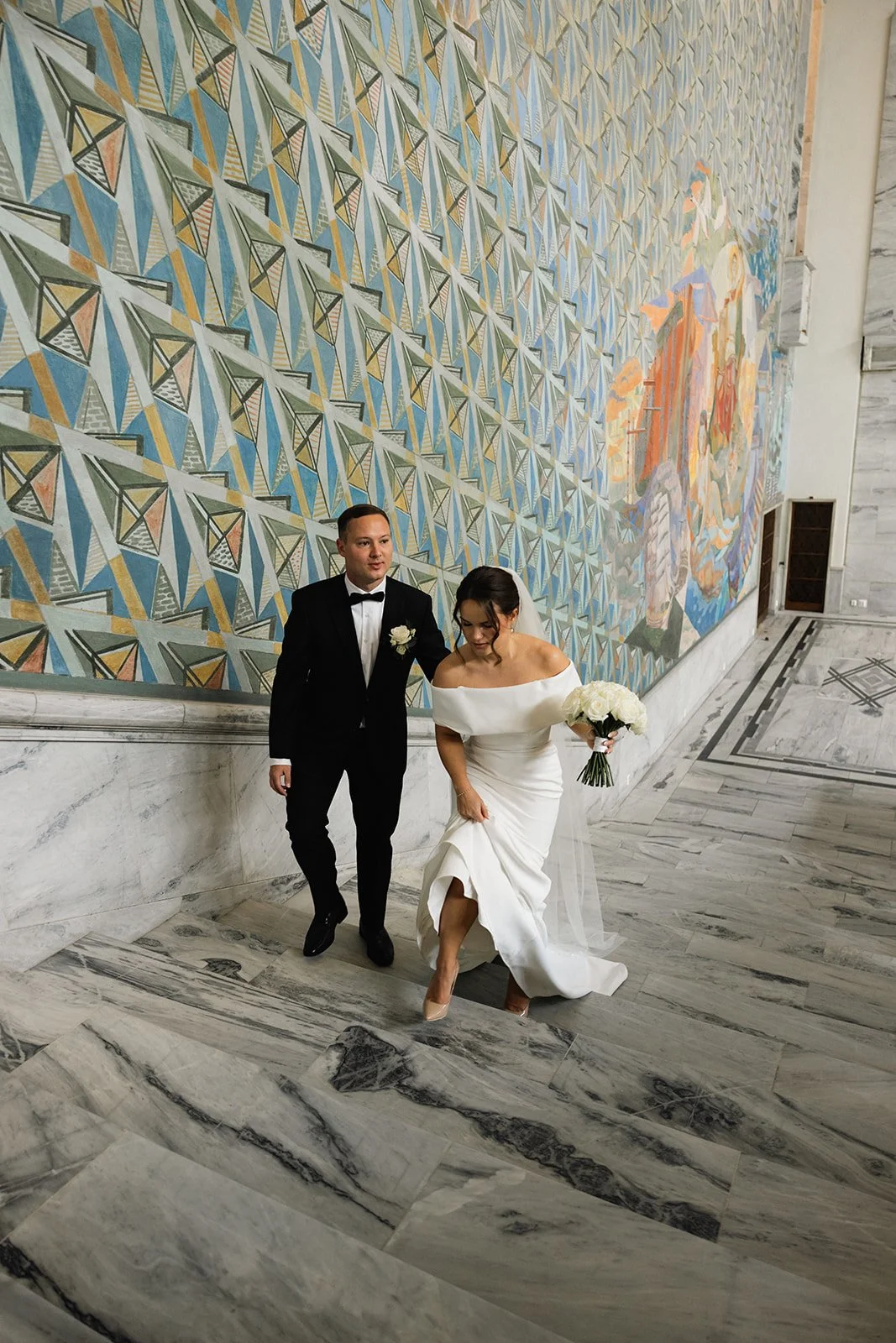 En brud og en brudgom på veg opp et marmortrapp, inne på et bygg med en stor, fargerik veggmaleri i bakgrunnen.
