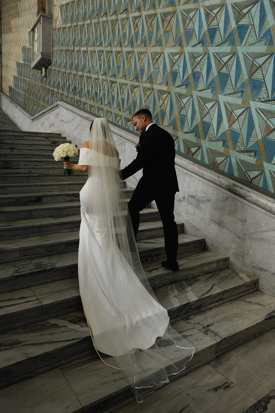 Et brudepar på en trapp, med bruden i en hvit kjole og lange skjørt og brudekjole, holder en blomsterbukett, mens brudgommen i en svart dress hjelper henne opp trappen, bakgrunn med fargerik geometrisk veggkunst.
