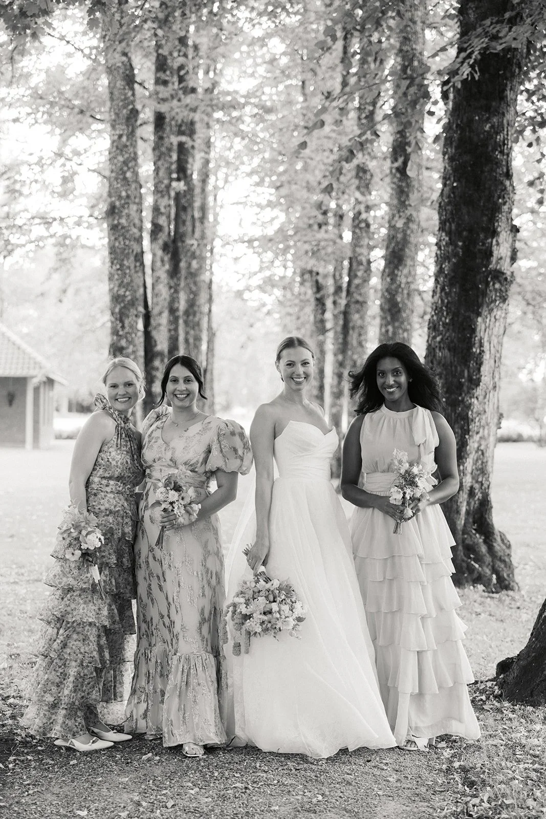 Fire kvinner i brudekjoler og blomsterbuketter står på hagekirkegård med trær i bakgrunnen, fotografert i svart-hvitt.