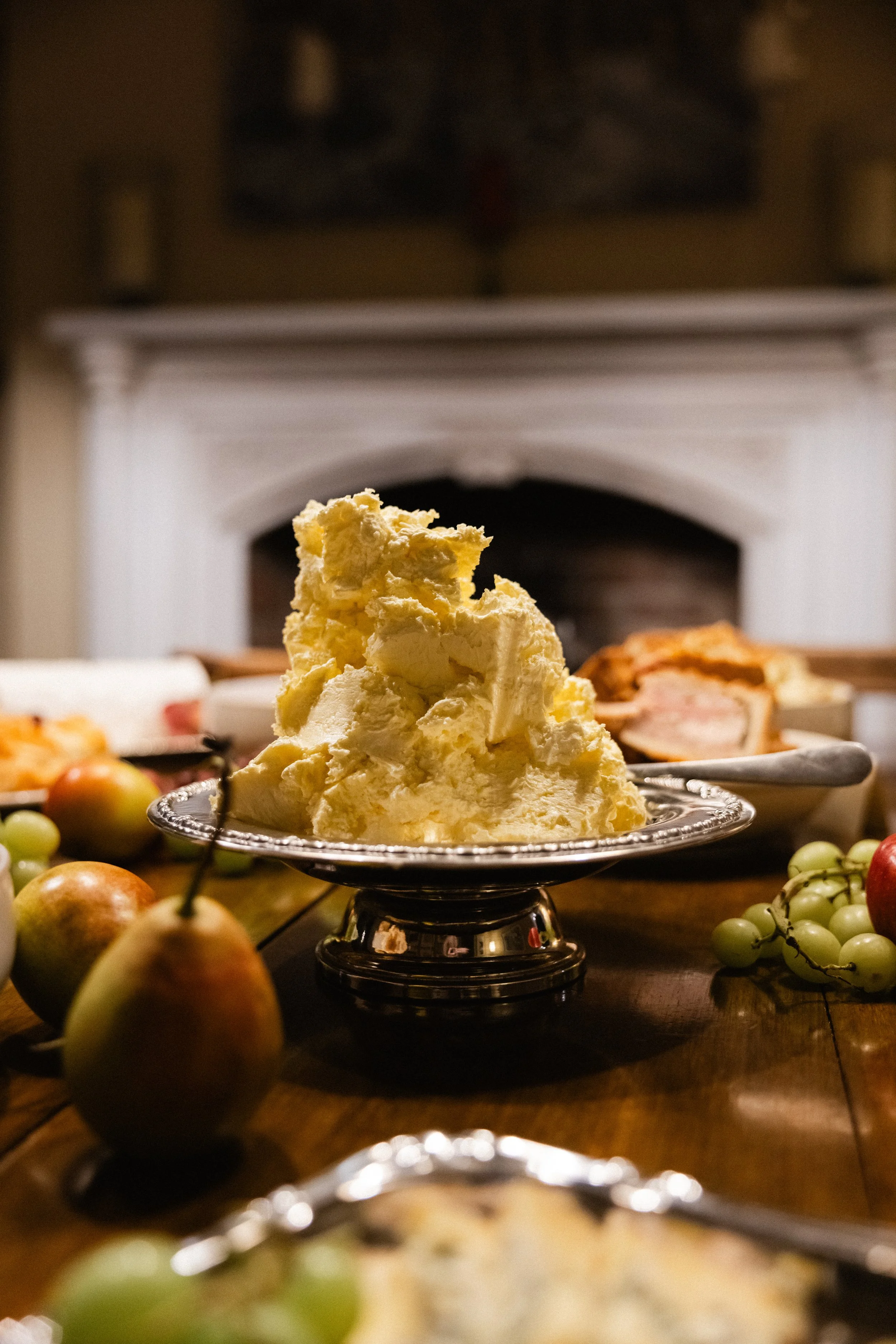 Et utvalg av ost på et sølvfat, omgitt av frukt som druer og pærer, med en bakgrunn av en peis.