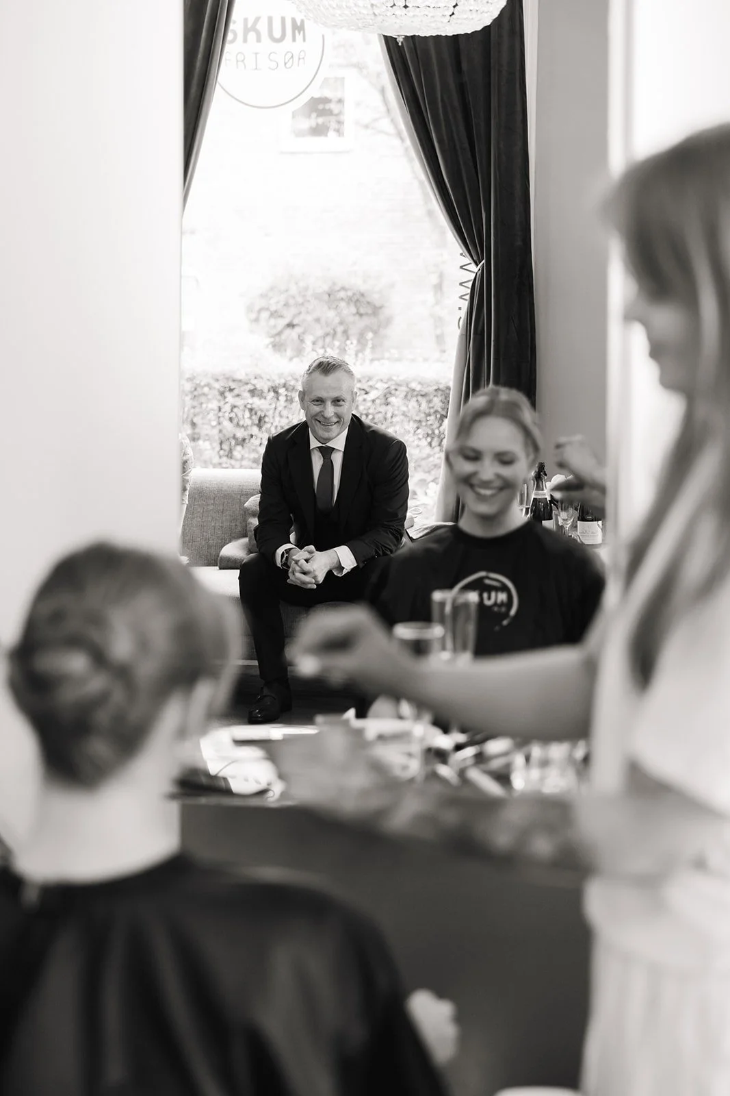 En gruppe mennesker sitter i et rom, der en person med ansiktsmaske og andre ser ut til å ha en samtale. Bakgrunnen viser et vindu med gardiner og et skilt som sier "SKUM FRISØR". Bildet er i svart-hvitt.