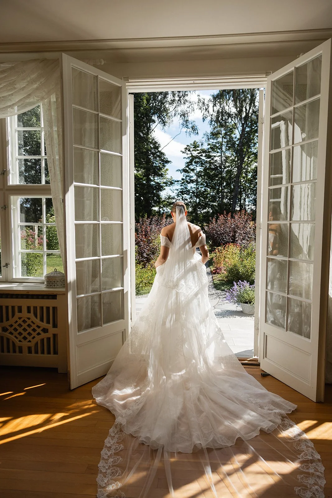 En brud står i en åpen dør som leder ut til en hage med blomster og trær på en solrik dag.