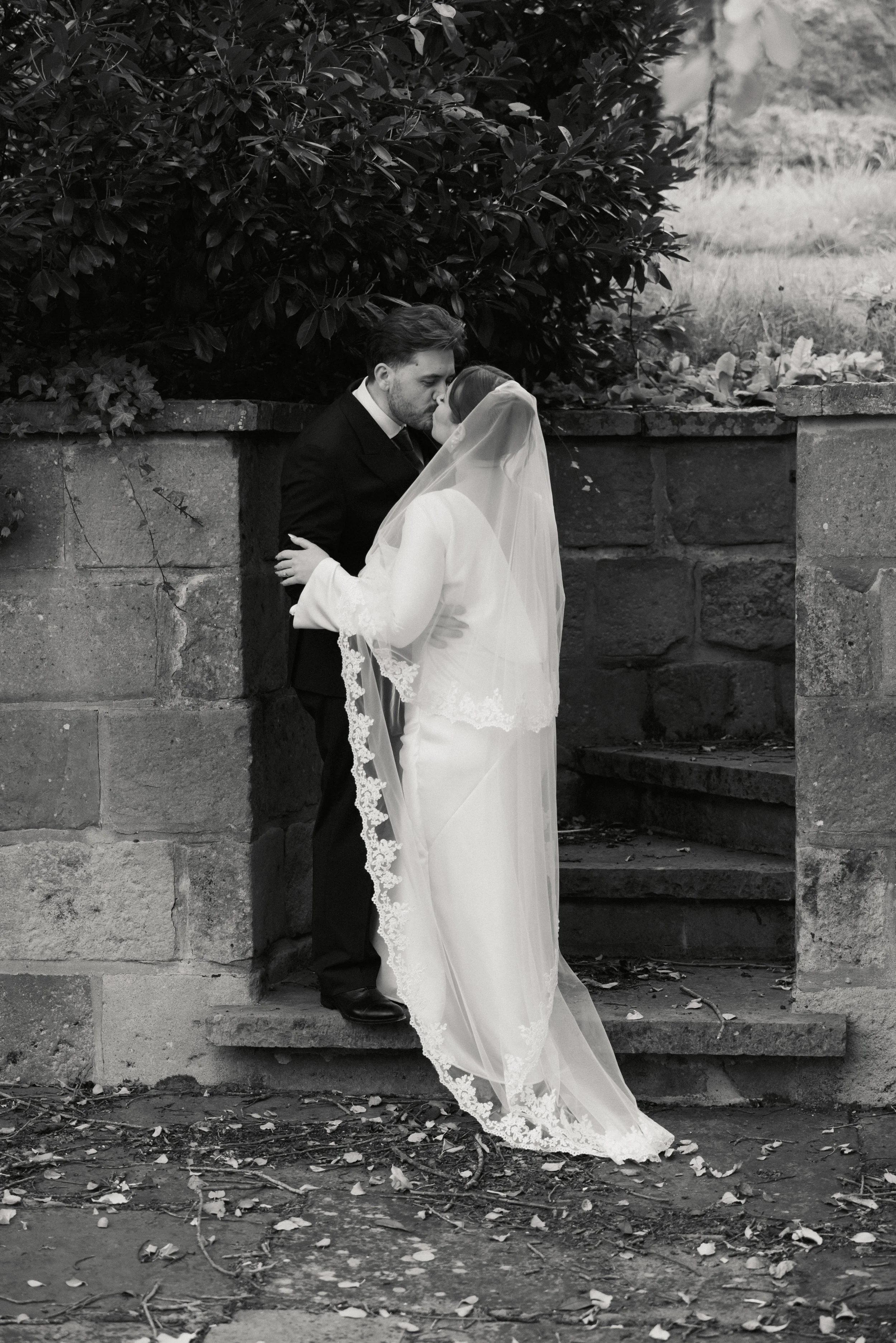 Et bryllupsbilde av et par som kysser, står på trappetrinn foran en steinmur med grønt løvverk i bakgrunnen.