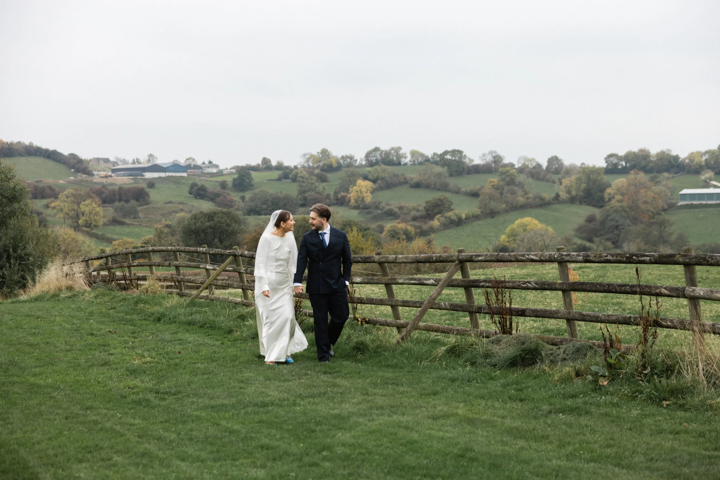 Et brudepar går hånd i hånd på en grønn eng med osteaktig tråkke, omgitt av bølgende grønne åser og spredte trær i bakgrunnen.