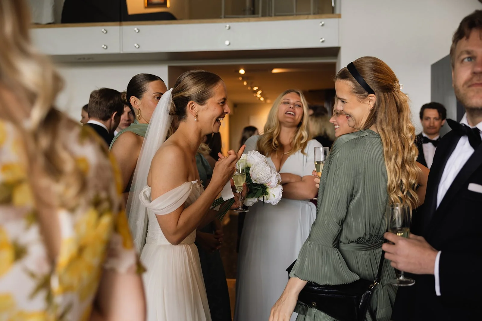 Måtefull gruppe mennesker som smiler og ler under en bryllupsfeiring, med bruden i fokus, som holder en blomsterbukett og har på seg en hvit bryllupskjole.