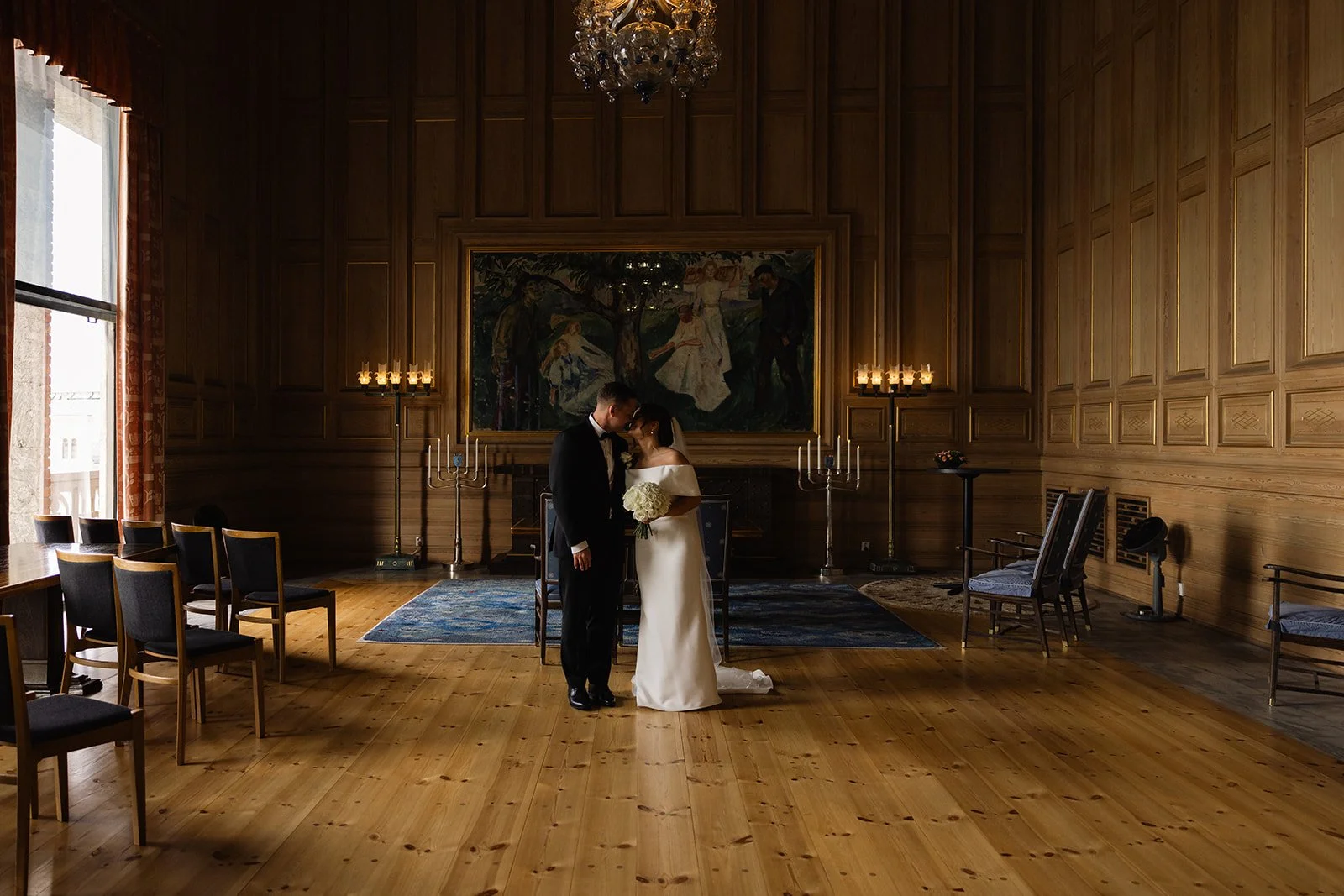 Et brudepar står sammen i en rom med tregulv og trevegger, omgitt av lysende lysestaker. De står foran et maleri i en lakonisk innramming, og det er stoler langs veggene.