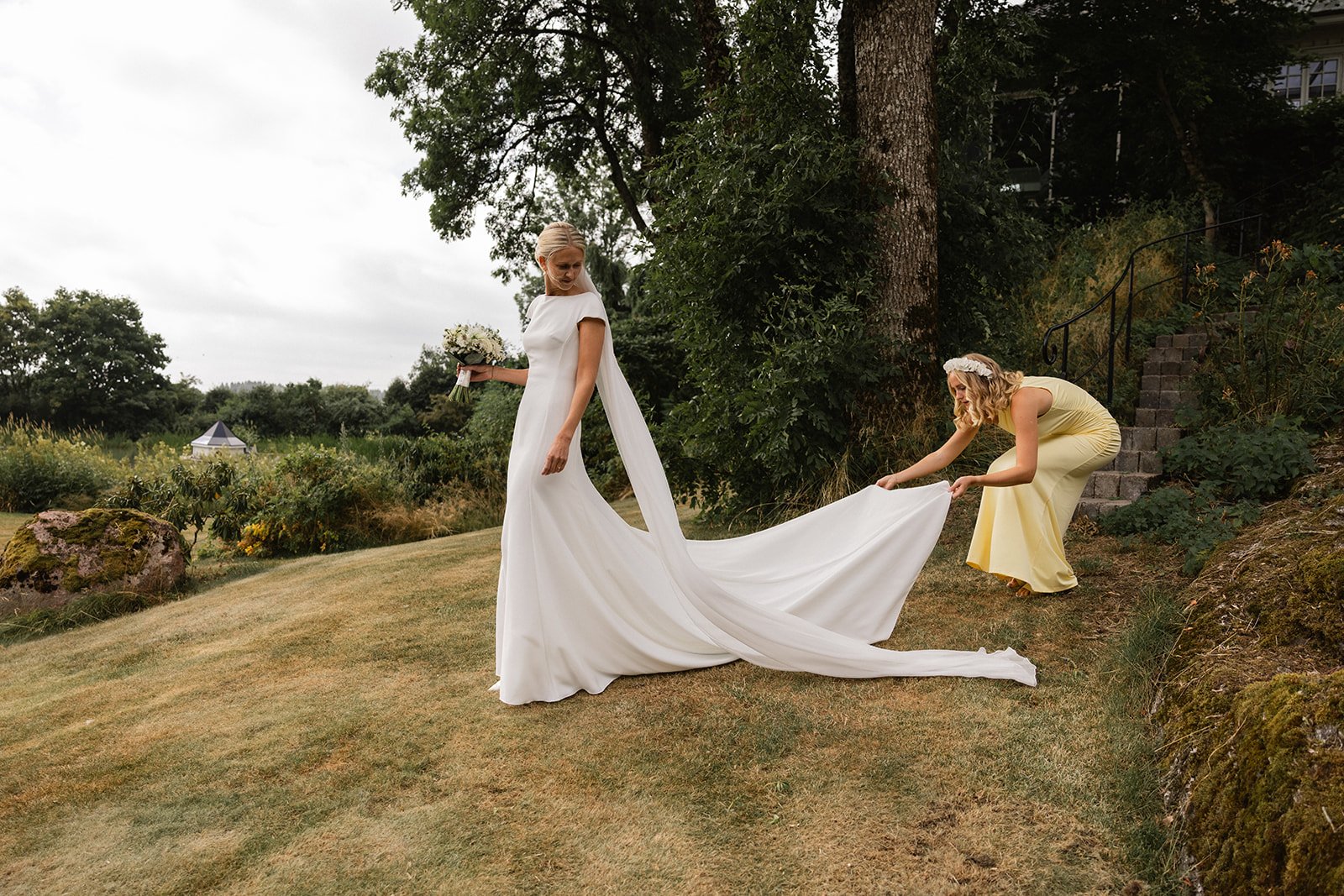 En brud i hvit kjole blir hjulpet av en annan kvinne i en gul kjole med å justere brudens lang hvite slør utendørs.