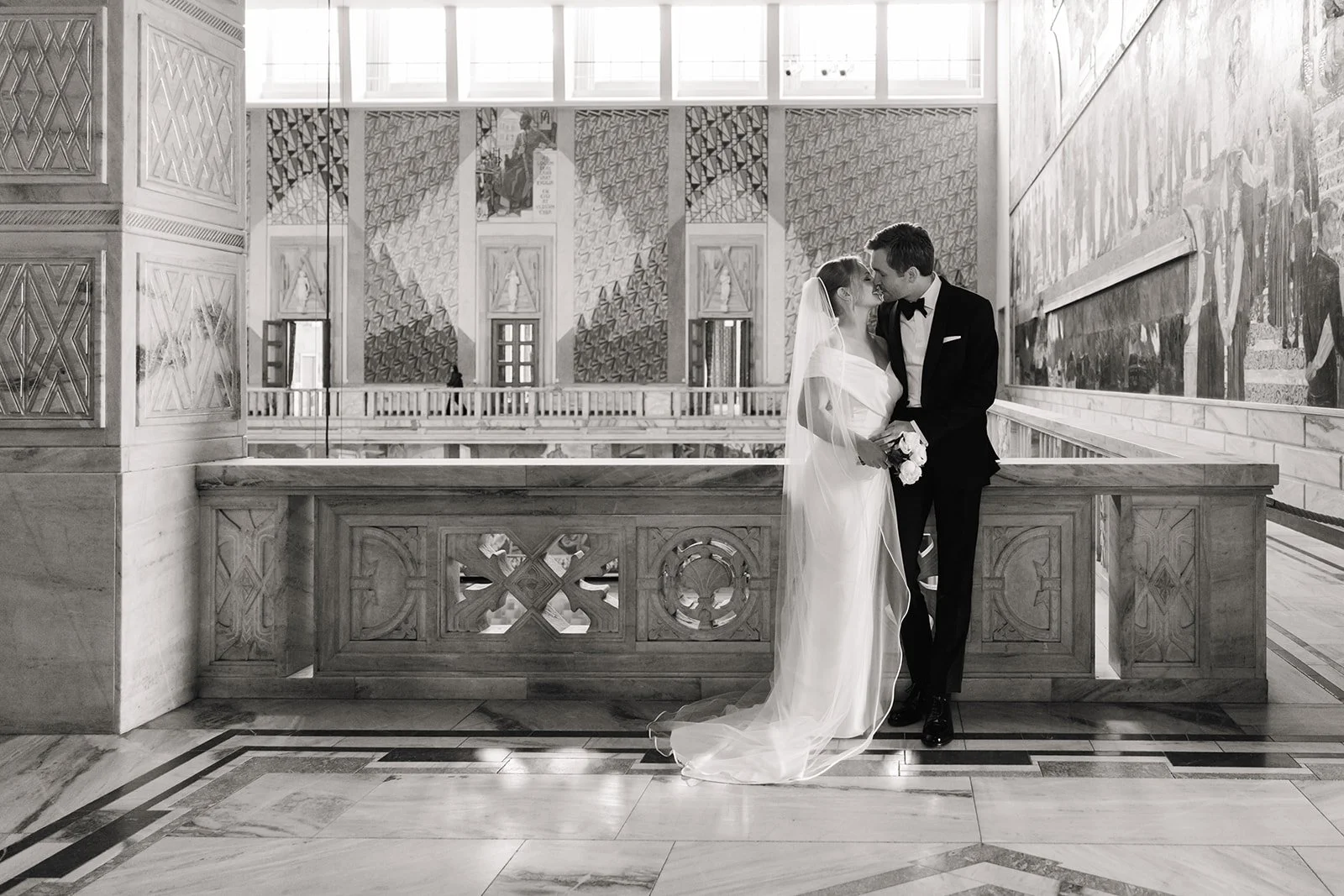 Et kjærestepar i bryllupsklær står og kysser i en elegant hall med kunstneriske veggdekorasjoner.