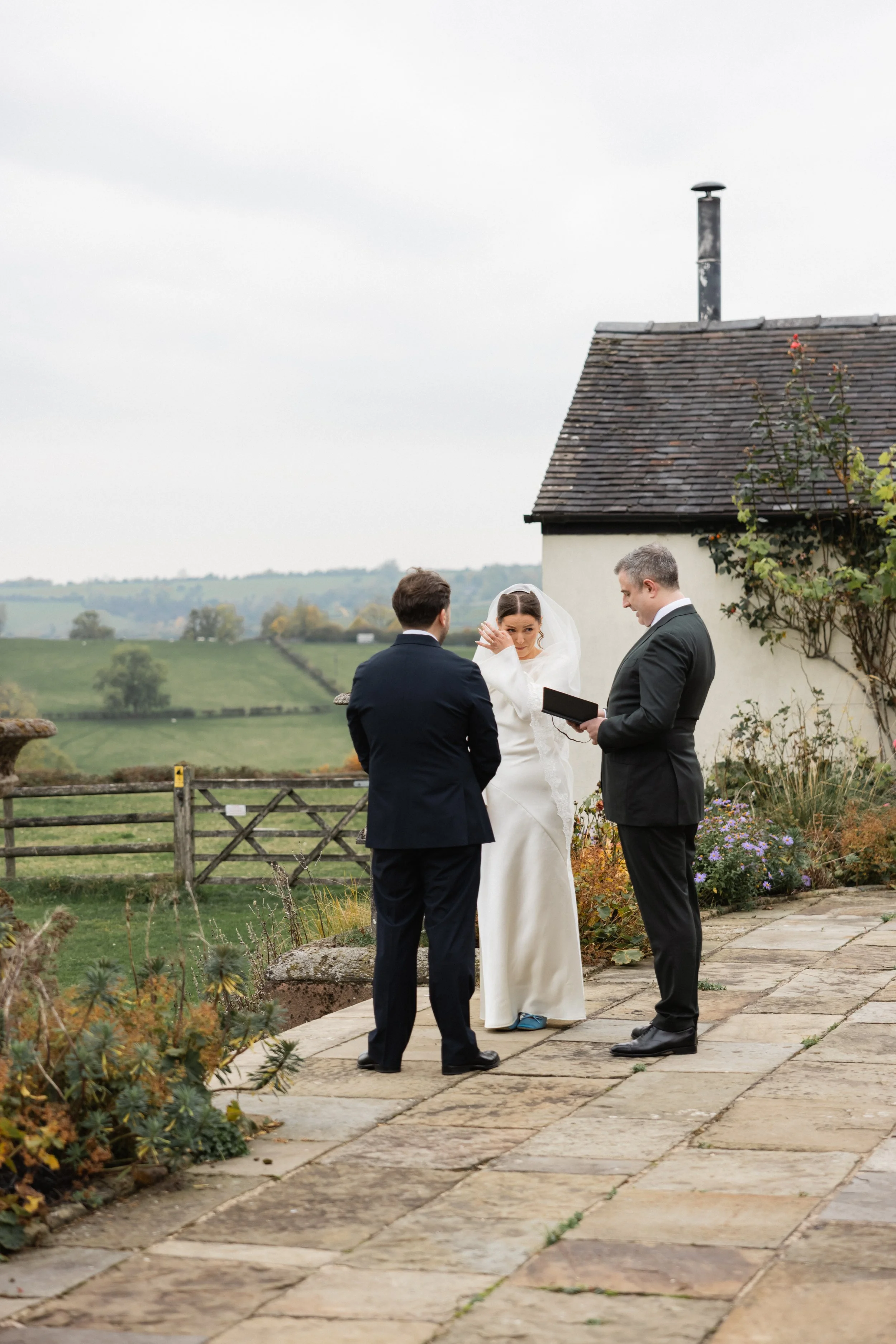 En bryllupsseremoni utendørs på en steinplattform med landskap i bakgrunnen, der en brud i hvit kjole og to menn i sorte dresser deltar i seremonien.