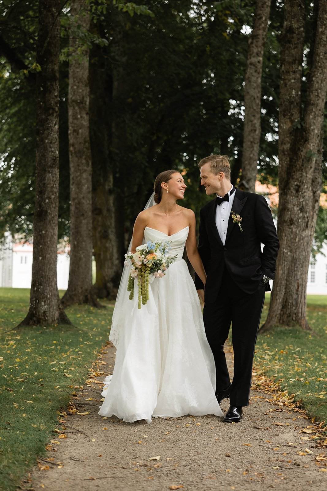 Et kledd brudepar går hånd i hånd langs en steinbane under trær. Bruden har på seg en hvit brudekjole og holder en blomsterbukett, mens brudgommen har på seg en svart dress med slips.