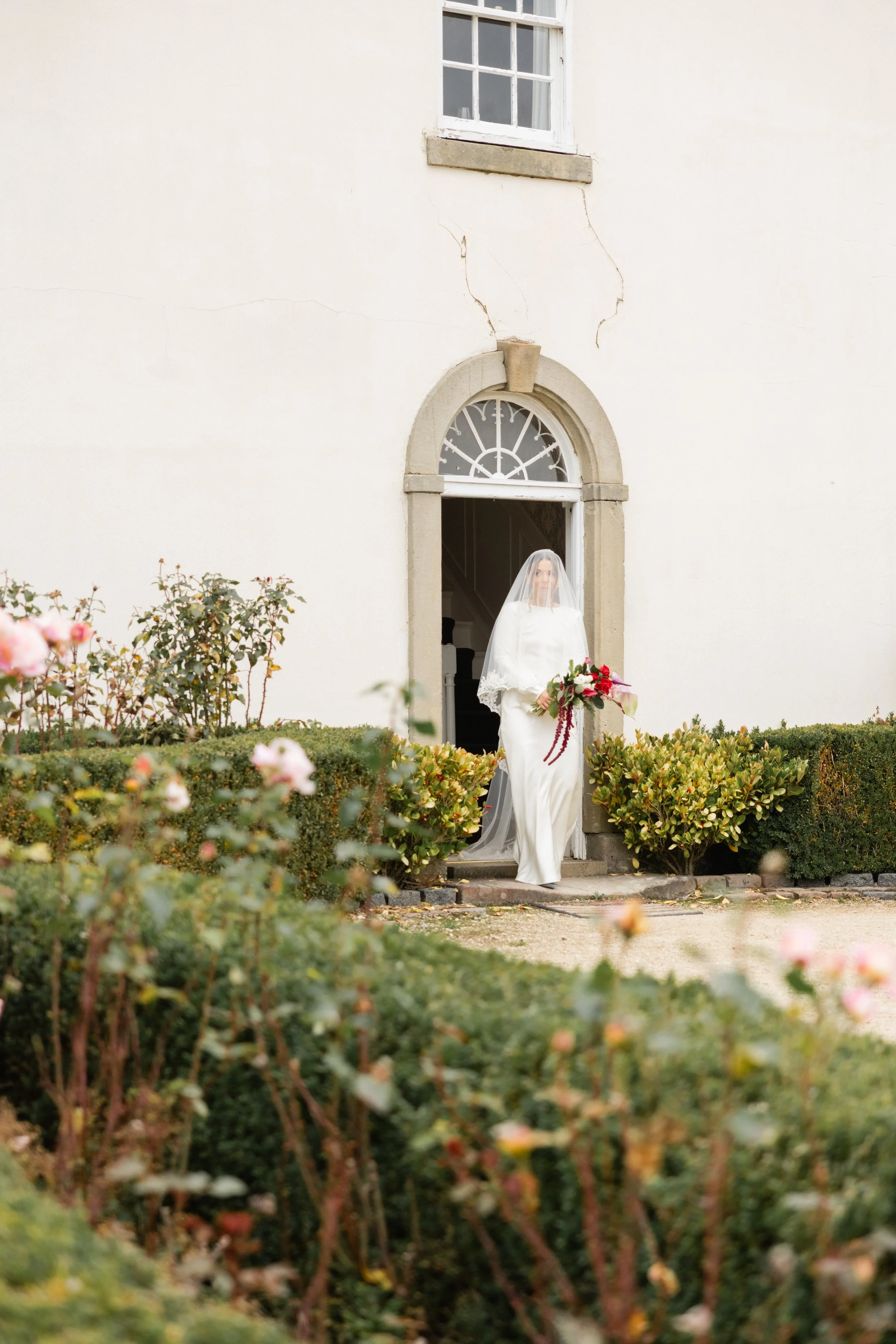 En brud i hvit brudekjole med slør som står i døren til en bygning, omgitt av grønne busker og blomster.