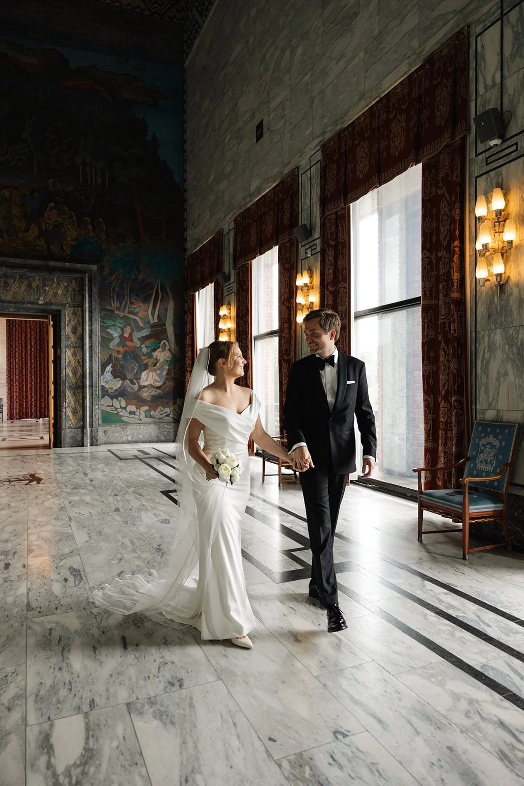 Et nygift par går hånd i hånd gjennom et elegant, lyst rom med store vinduer, marmorgulv og dekorative gardiner, smilende og kledd i bryllupsantrekk.