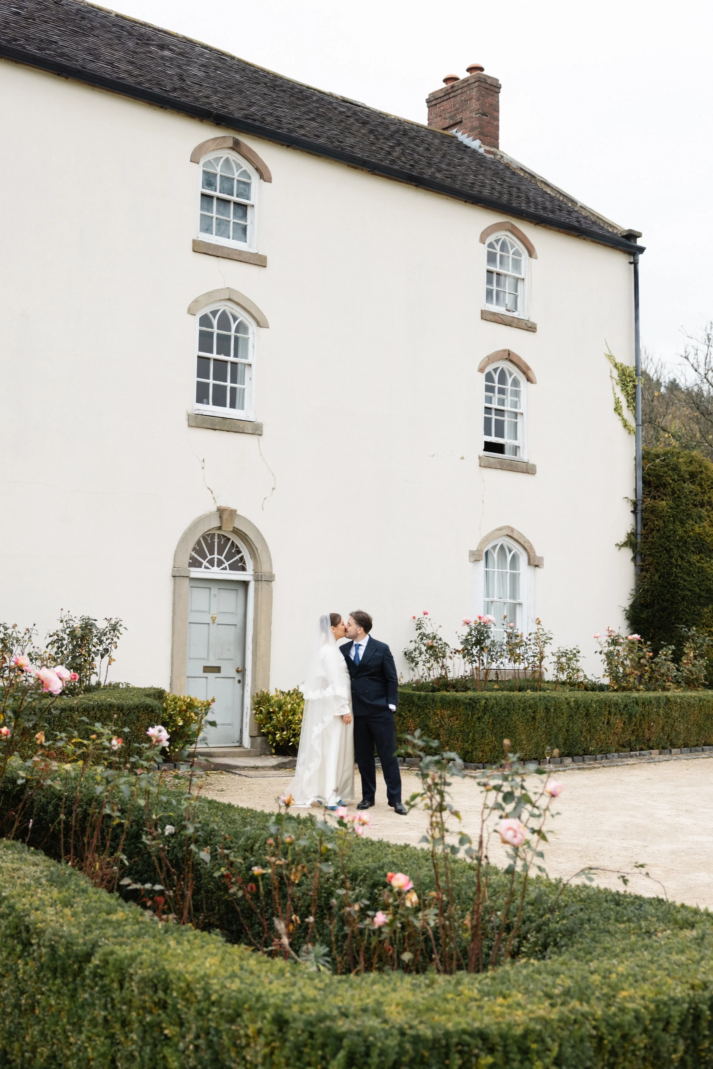 Et brudepar som kysses foran en hvit murbygning med flere vinduer og en dør, omgitt av hekk og blomster i en hage.