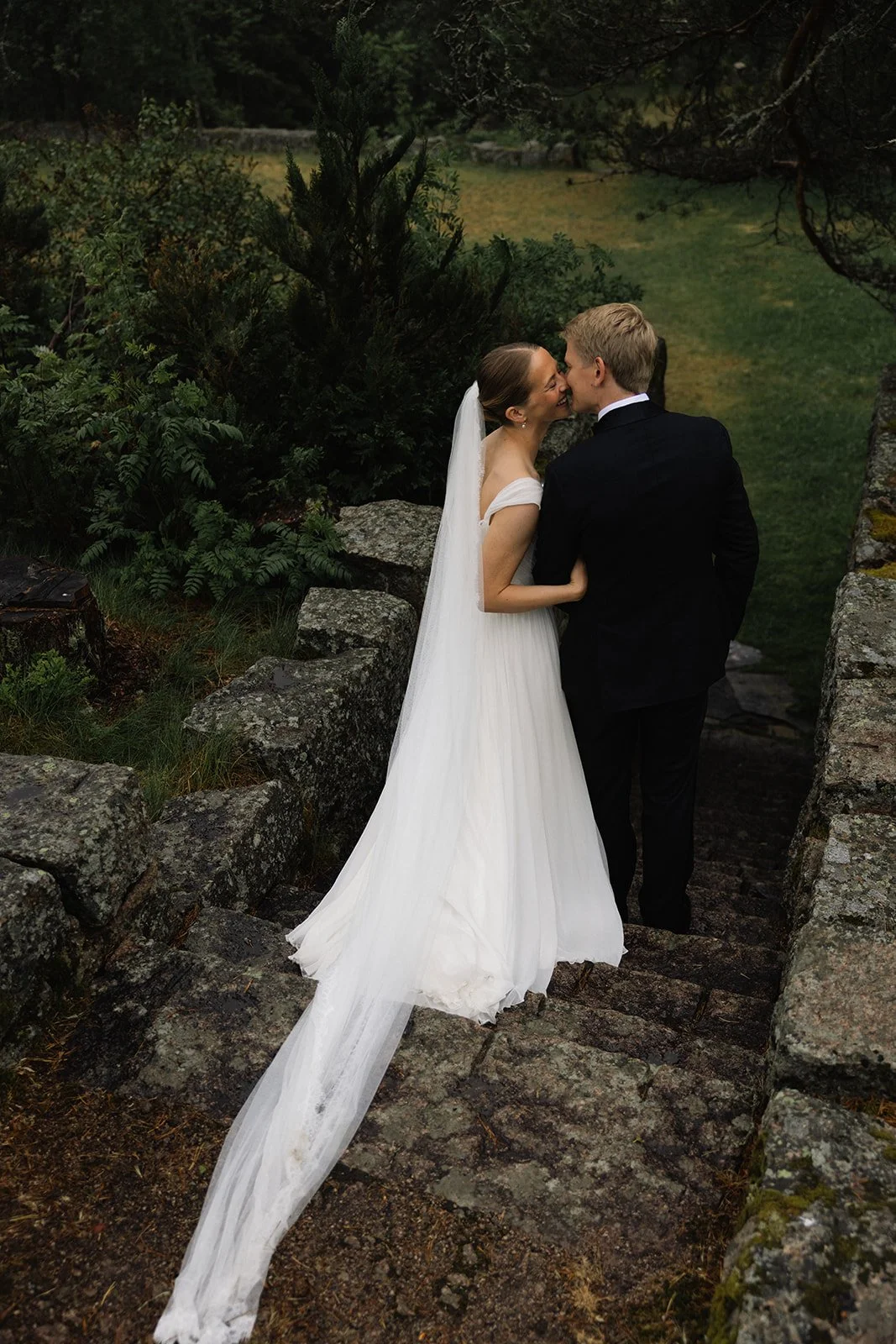 Nydelig brudepar som kysser utendørs, omgitt av natur og gamle steinmurer.