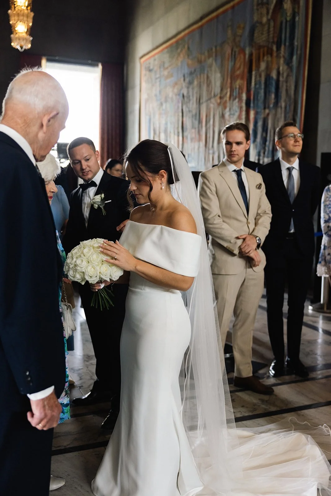 En brud i en hvit kjole holder en bukett med hvite roser under et bryllupsseremoni, omgitt av flere kleskledde gjester.