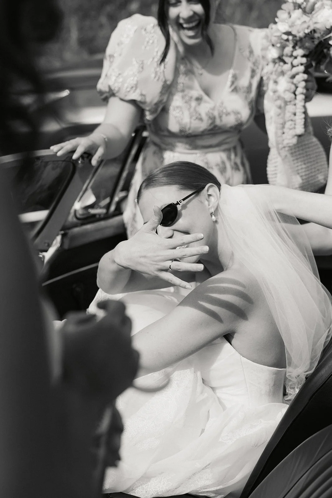 En brud med mørkt, rett hår og høye øredobber, iført brudekjole og veil, sitter i en bil og dekker ansiktet med hånden, mens en annen kvinne ler i bakgrunnen.