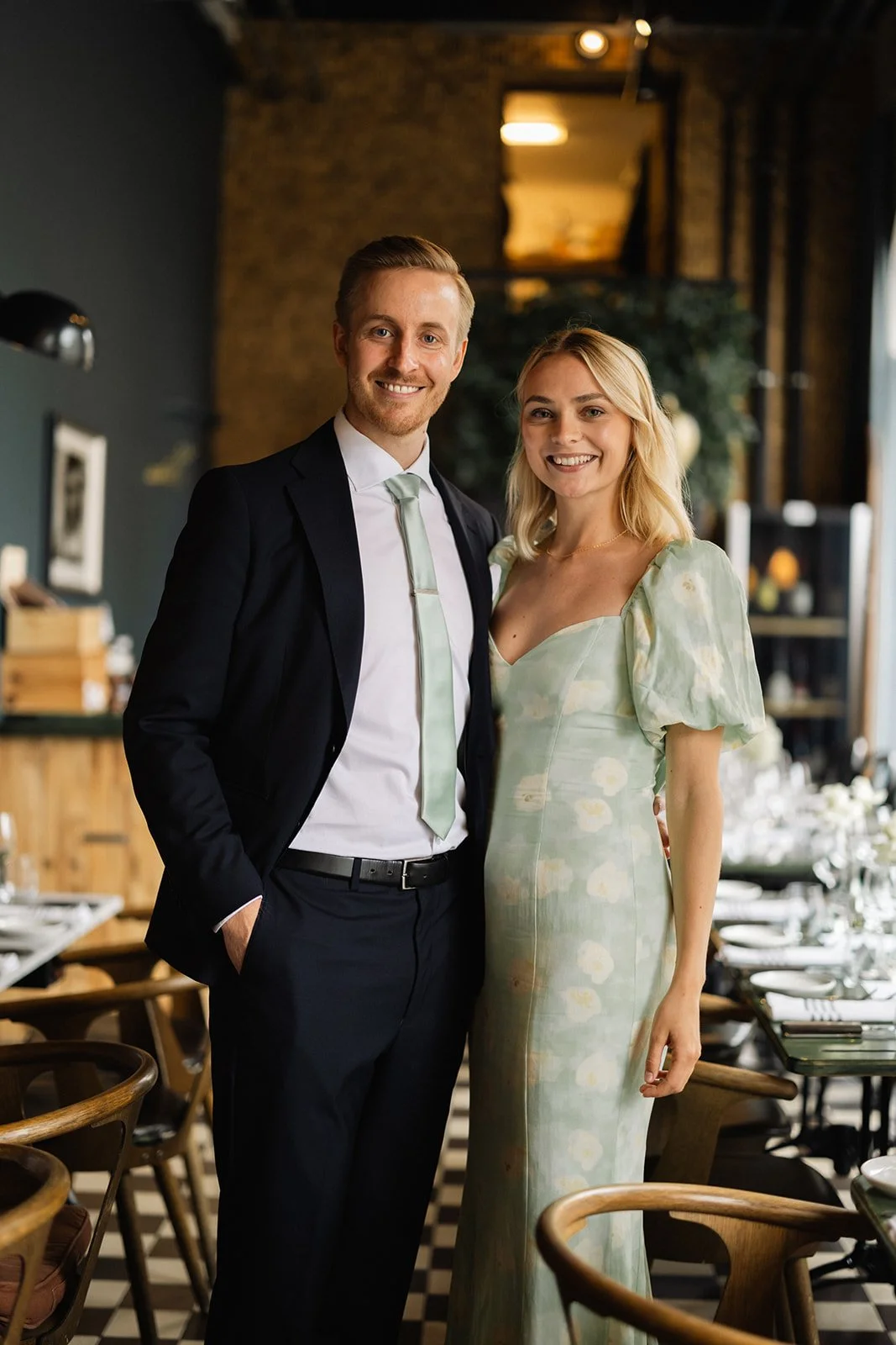 En man og en kvinne smiler og poserer sammen i et innendørs miljø, med et spisebord og stoler i bakgrunnen.