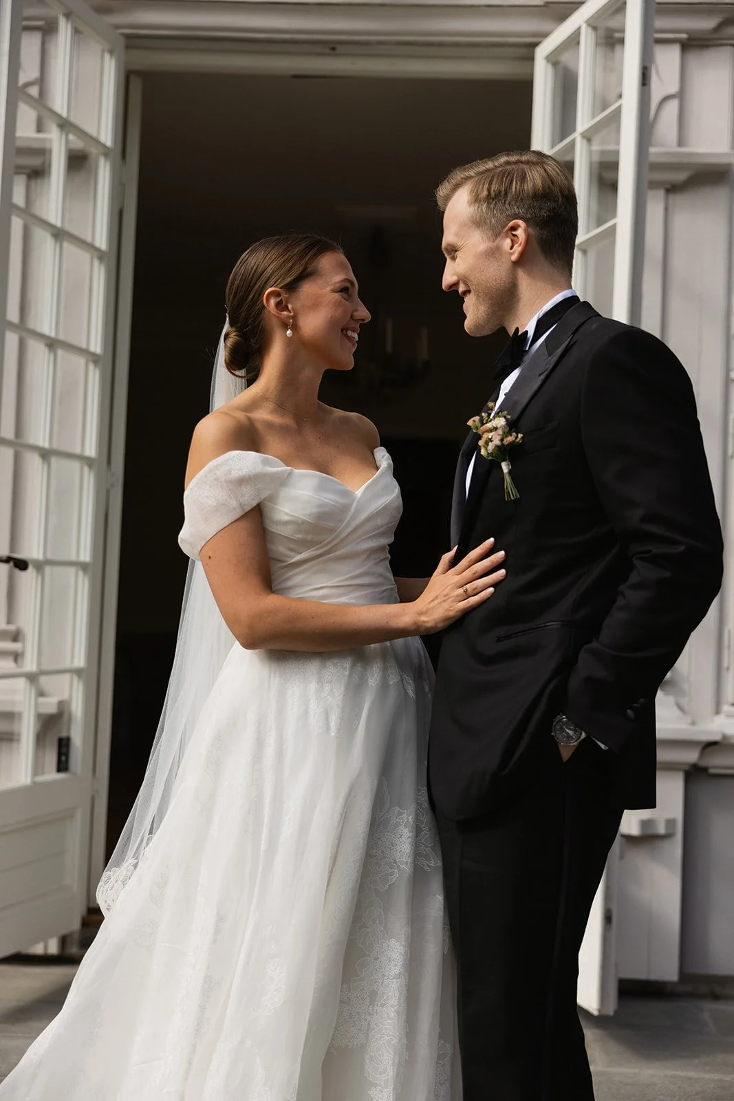 Et brudepar står inntil hverandre, smiler og ser inn i øynene, foran en åpen dør med hvite rammer.