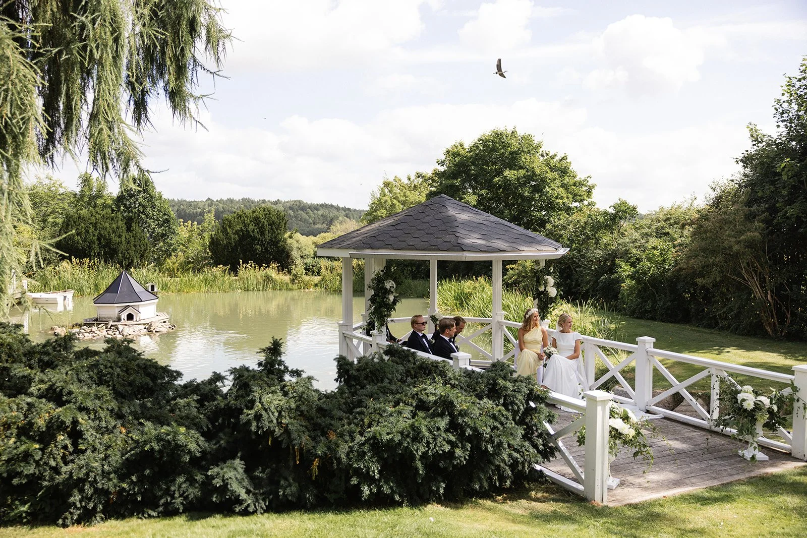 Et brudepar og fire andre sitter i en lystig paviljong ved en innsjø, omgitt av grønt landskap, der det er en liten øy med en liten bygning og en hekk.