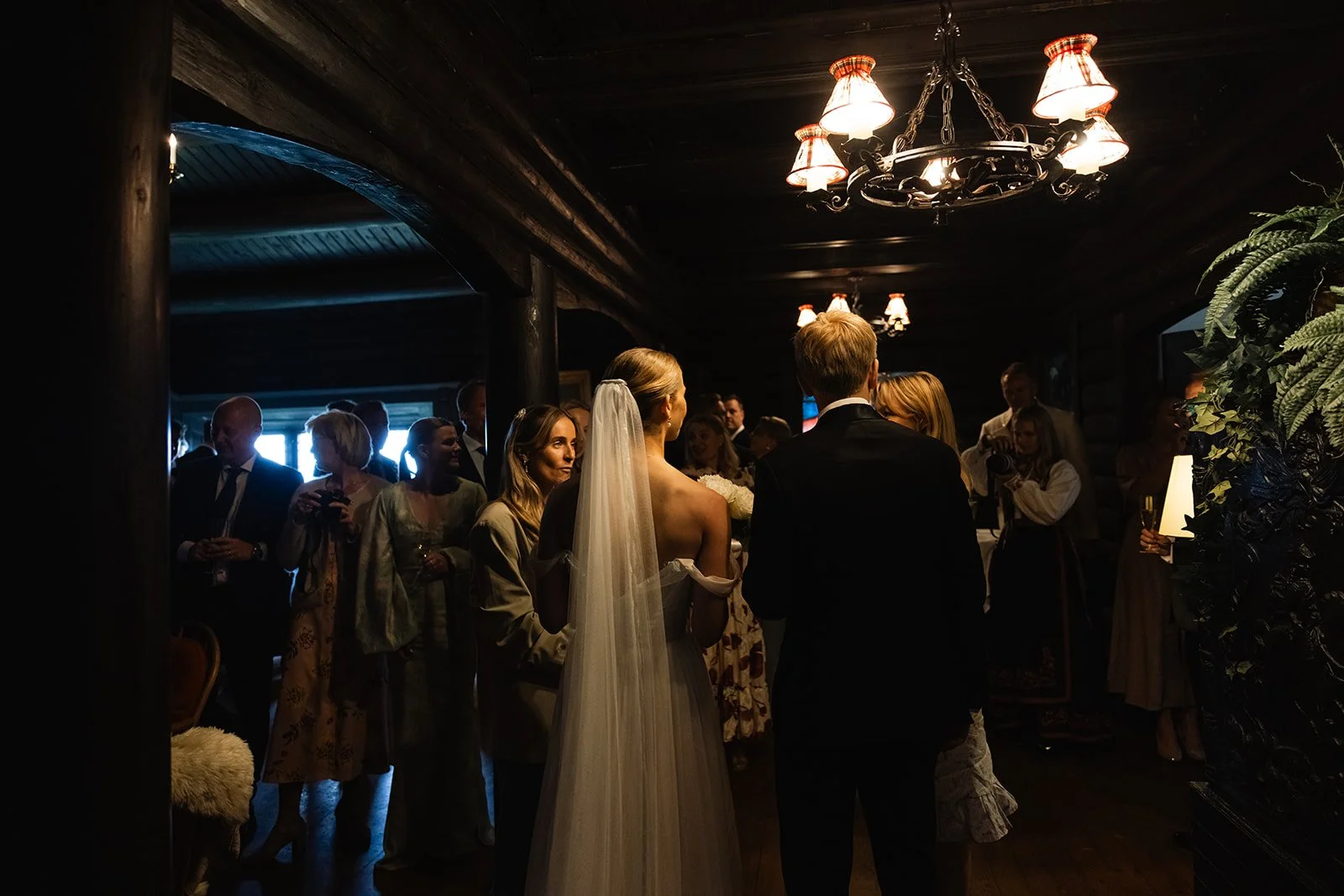 Bryllupsscene med bruden og brudgommen i midten, omgitt av gjester i et mørkt rom med vintage lysekroner