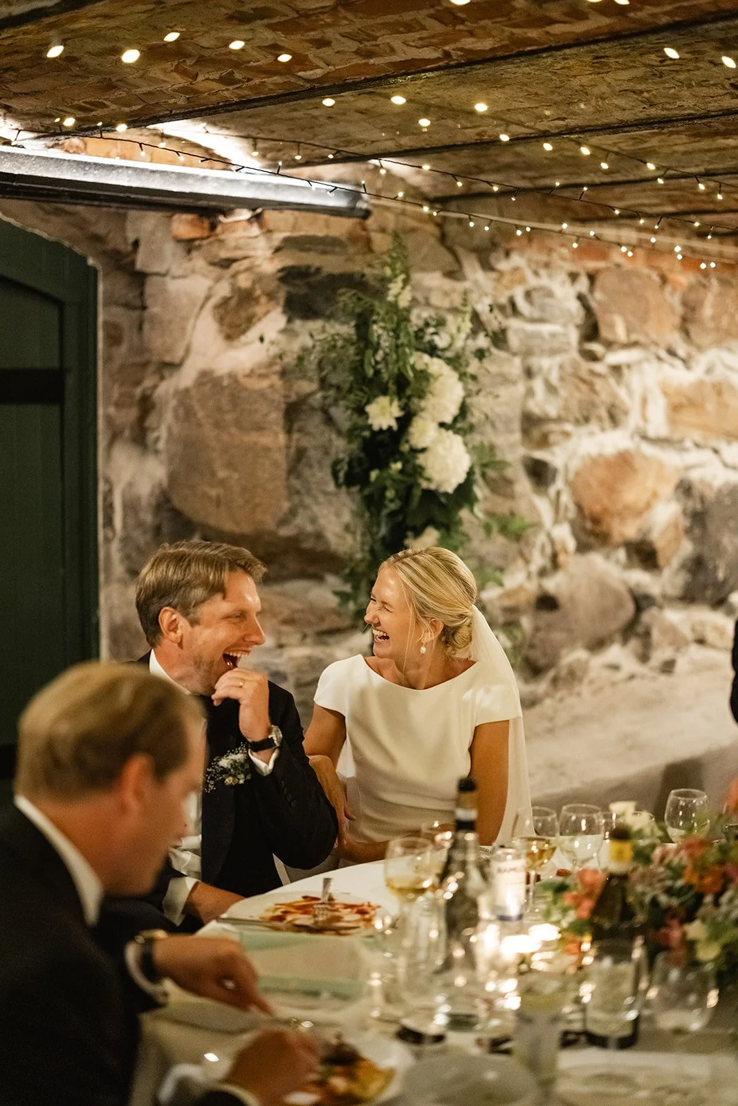 Mann og kvinne som ler og snakker under en fest, med mat og drikke på bordet, innendørs dekorert med blomster og lys