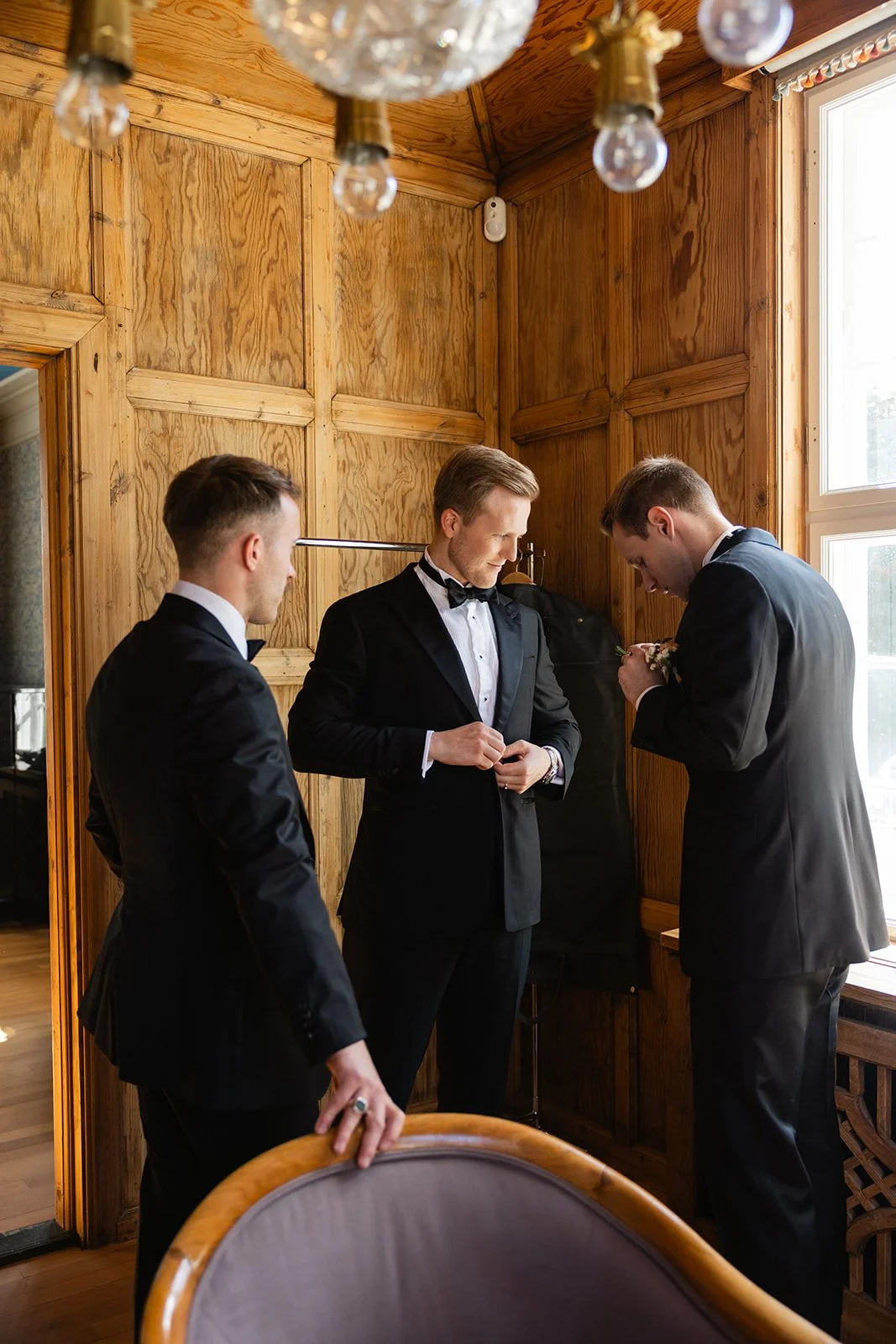 Tre menn i formelle klær, som ser ut til å være i en bryllupsforberedelse, står inne i et rom med trepanel og store vinduer.
